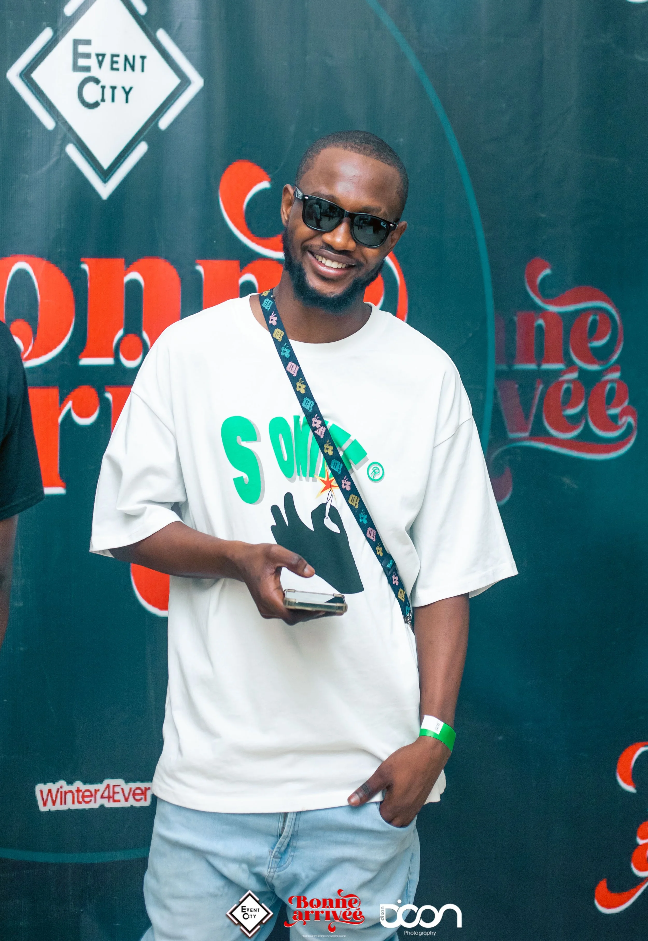 A young man smiling, wearing sunglasses and a white T-shirt with colorful text and graphic, holding a phone, standing in front of a backdrop with logos and text for an event.