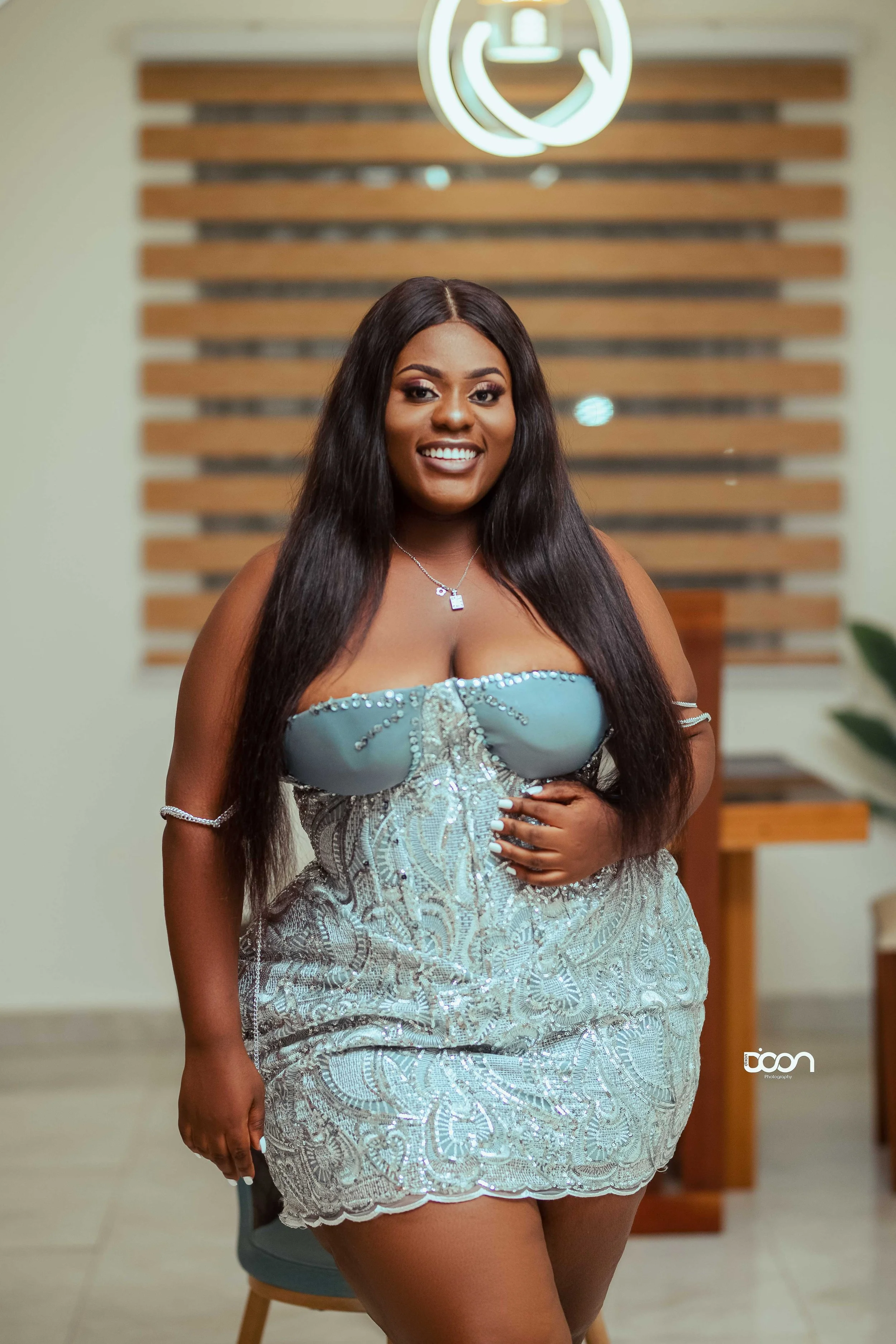 A smiling woman with long dark hair in a strapless silver dress stands indoors, in front of a wooden feature wall with a logo illuminated above her head.