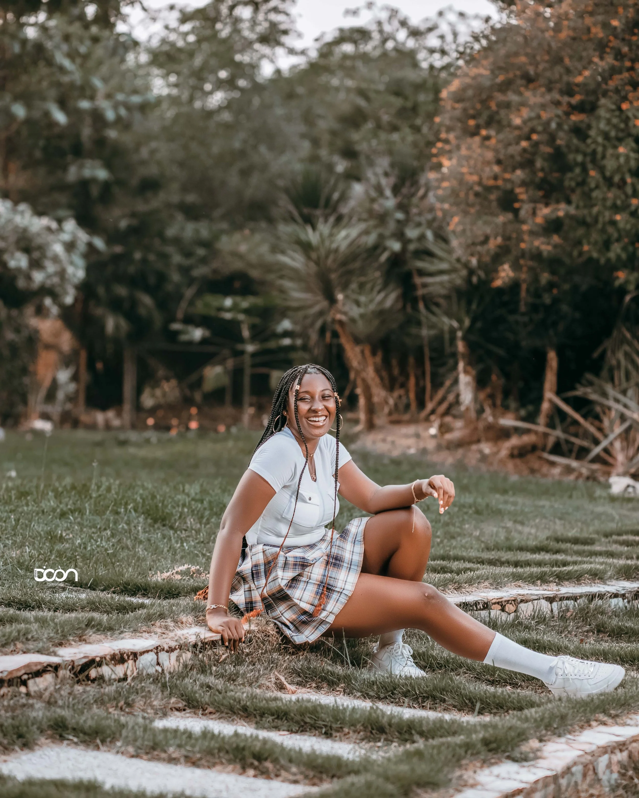 A young woman with braided hair, smiling and sitting outdoors on grass in a park-like setting with trees in the background, wearing a white t-shirt, plaid skirt, white socks, and sneakers.