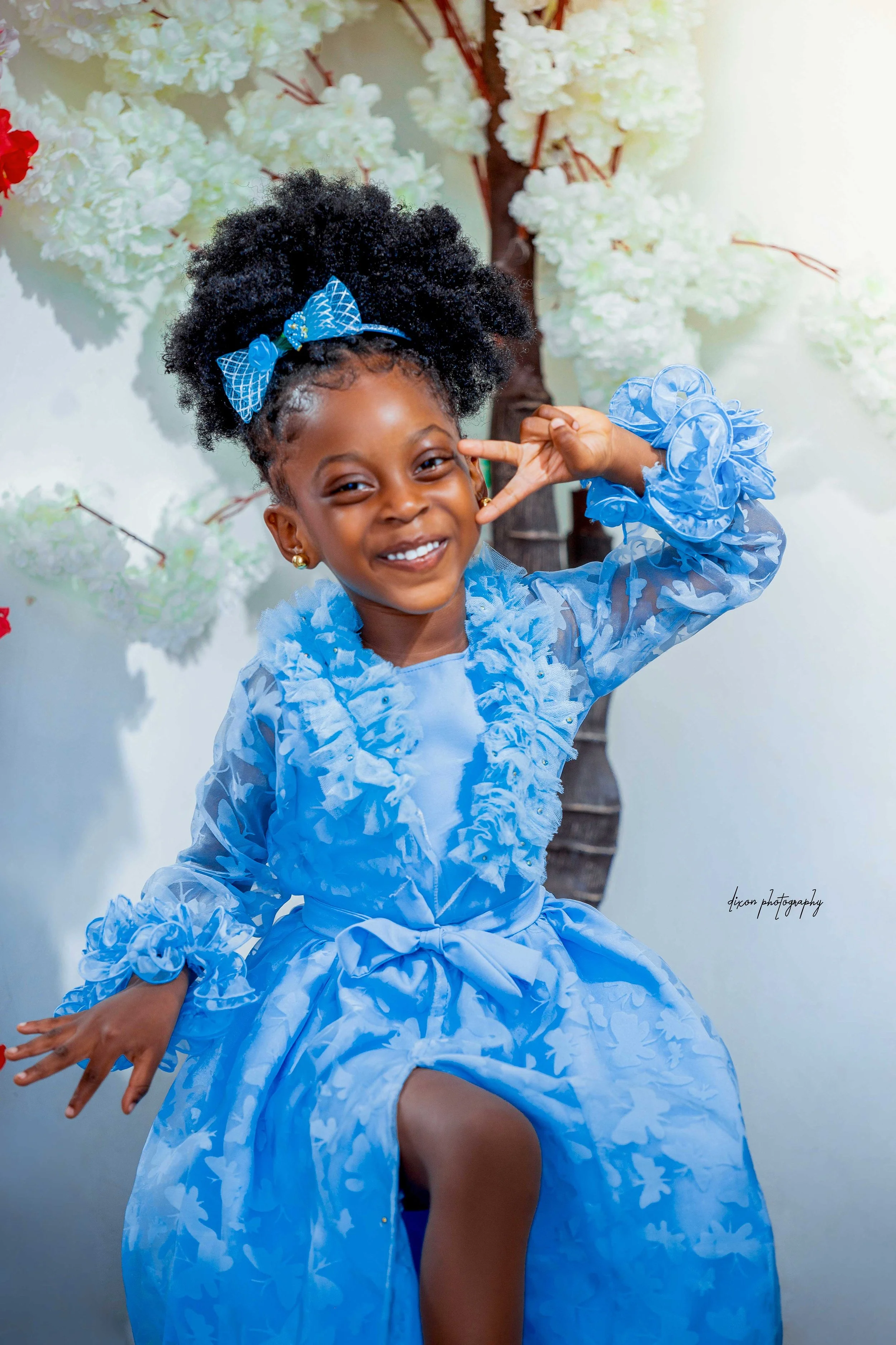A young girl wearing a bright blue dress with ruffled details and a matching bow in her hair, smiling and posing playfully with her hand near her face, standing in front of a background decorated with white flowers.