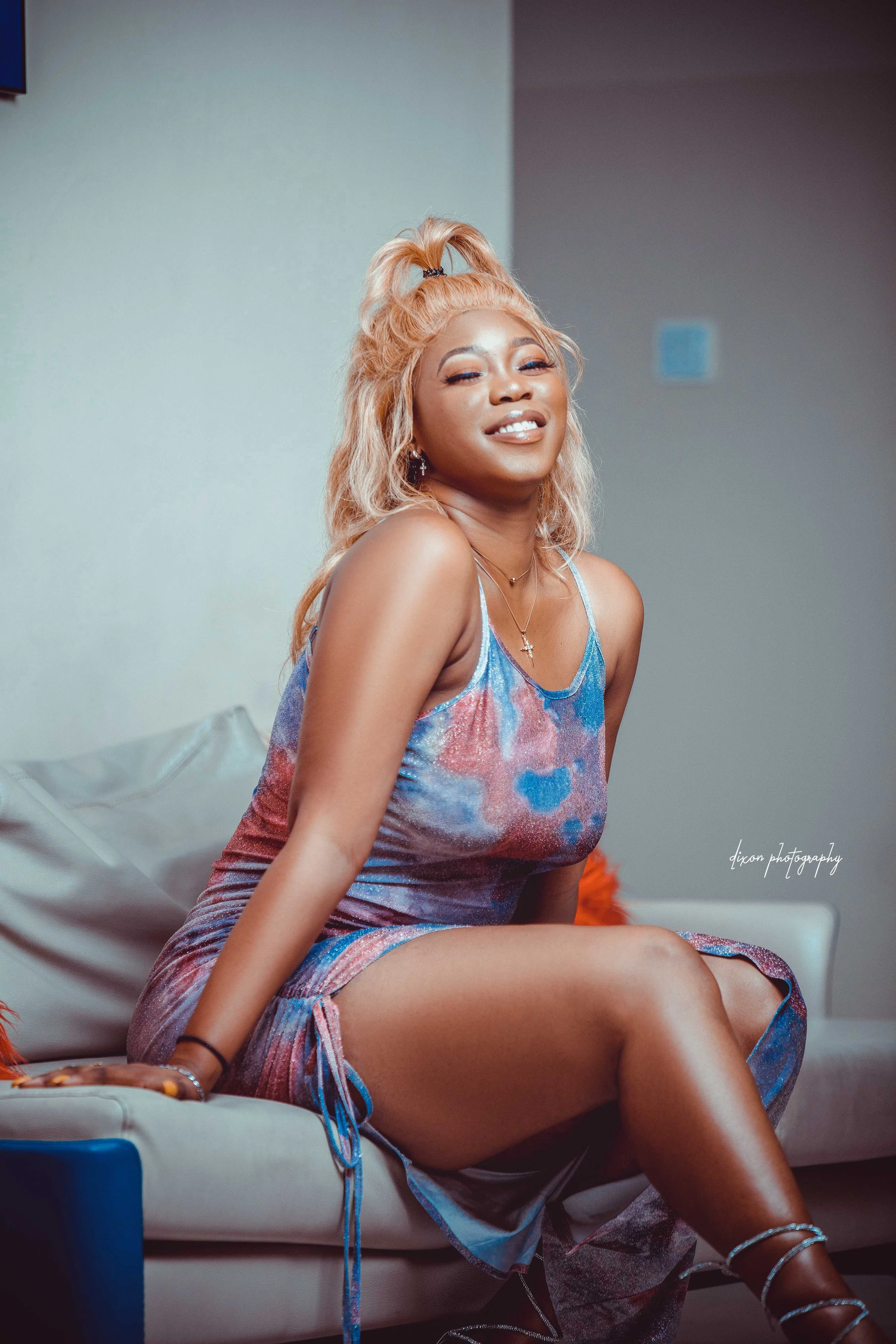 A young woman with blonde hair styled in a high ponytail, sitting on a beige couch, wearing a colorful sleeveless dress, and smiling at the camera.