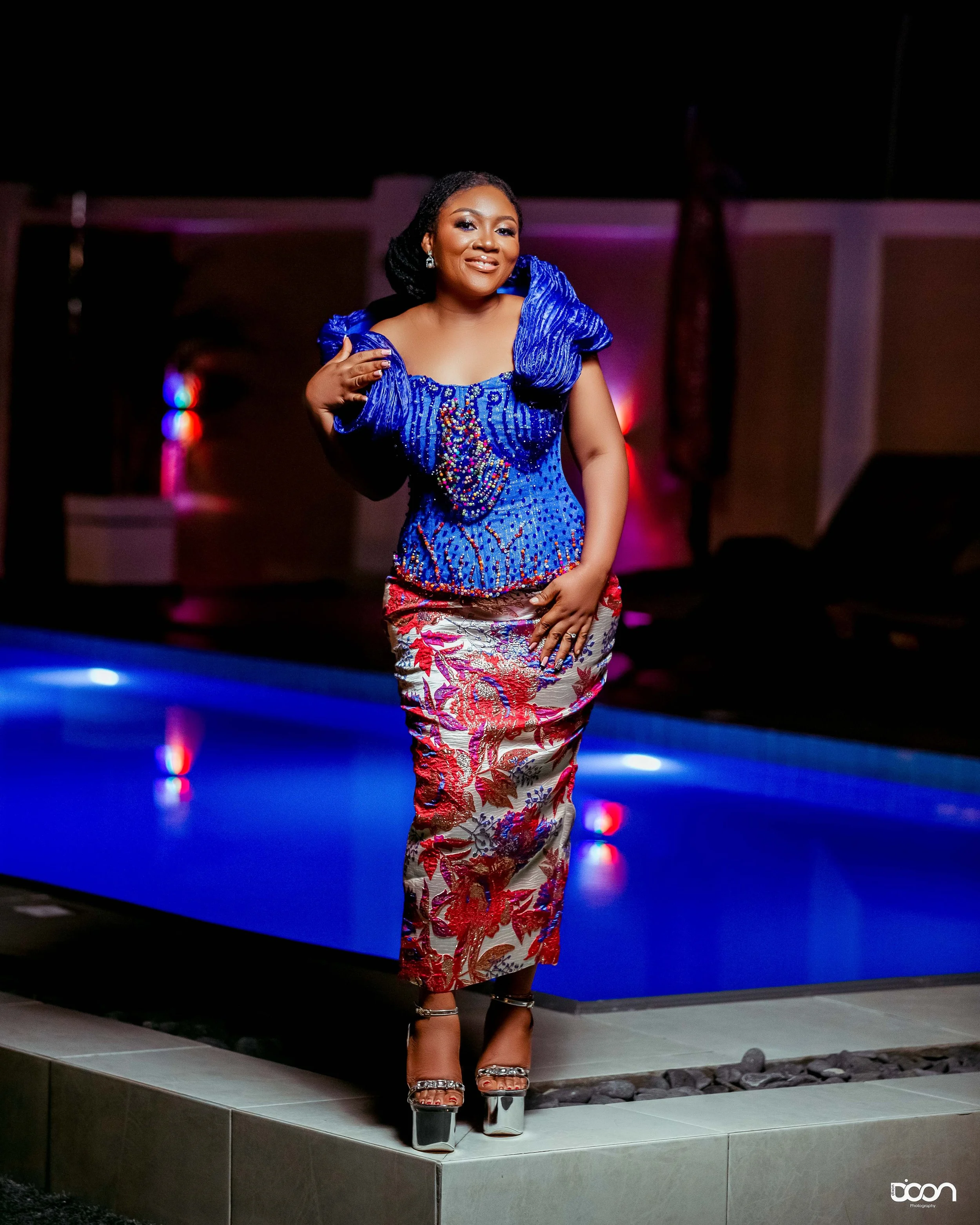 Woman in a vibrant blue top and patterned skirt standing near a pool at night, smiling and posing for the camera with colorful lighting in the background.