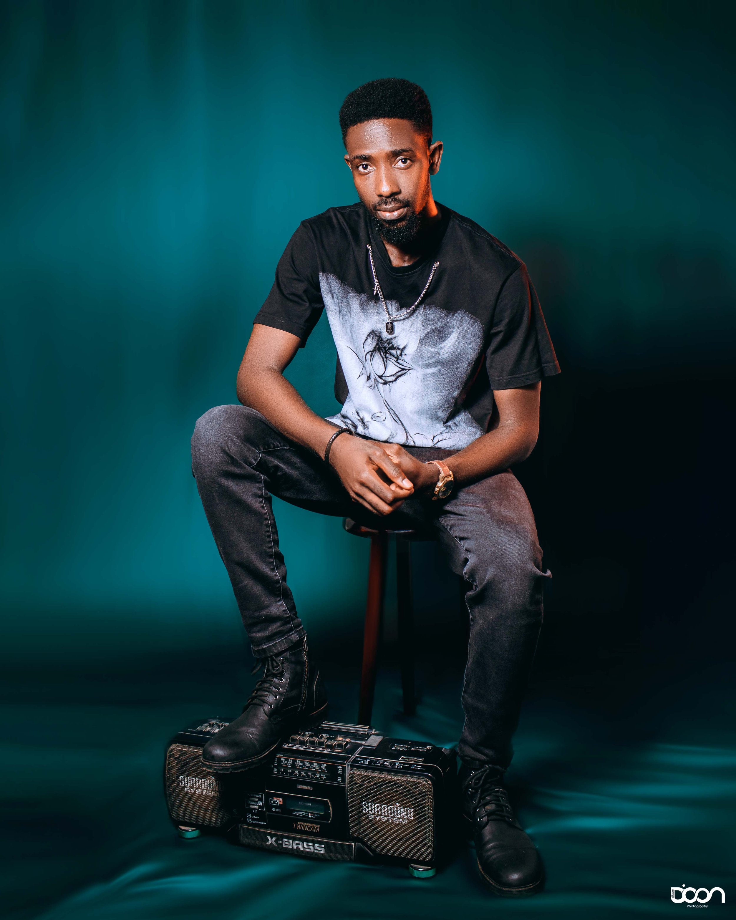 A young man with dark skin, short curly hair, and a beard, sitting on a stool with one foot resting on a vintage boombox, dressed in a black T-shirt with a white graphic, black jeans, and black boots, against a dark teal background.