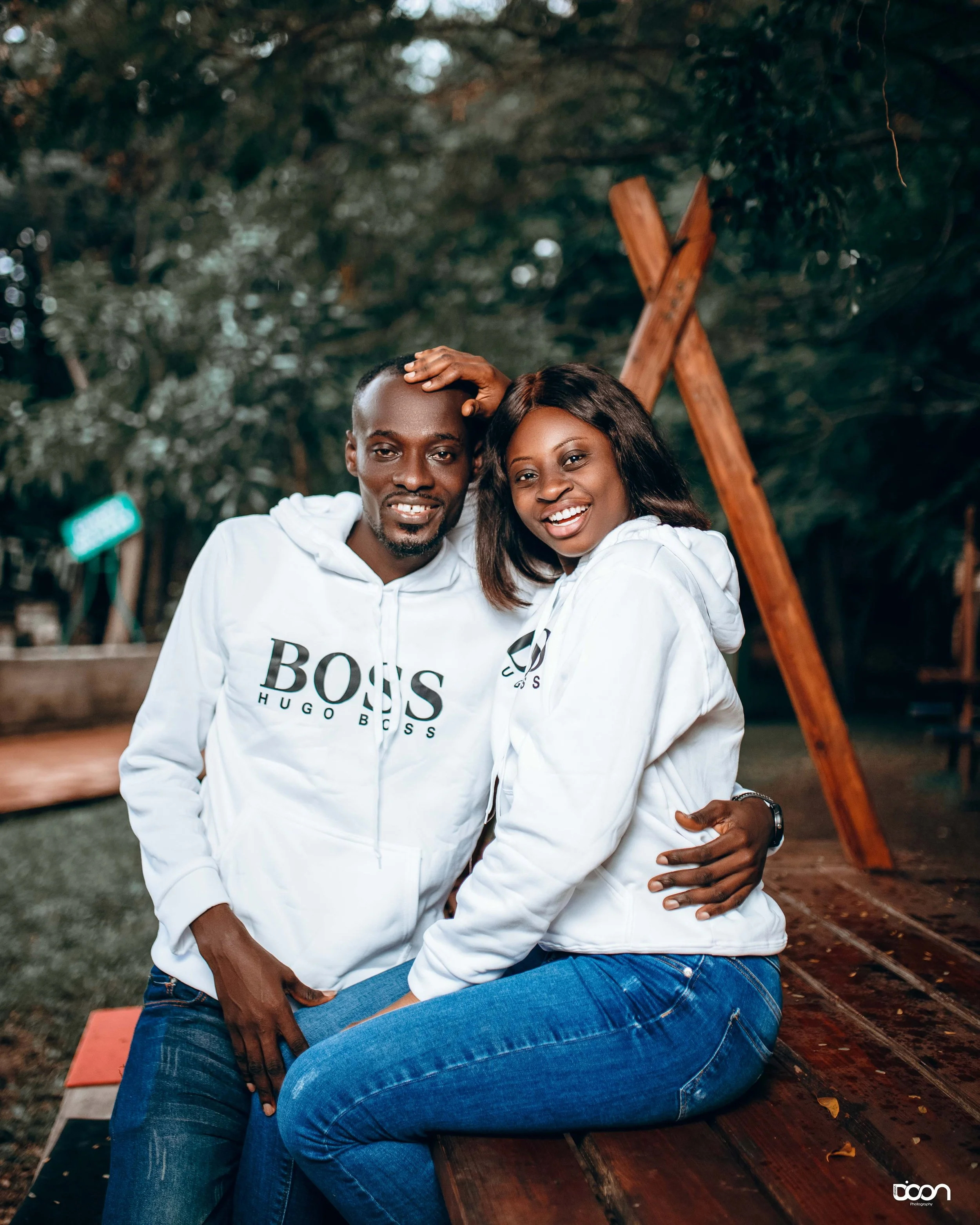 A smiling black couple sitting on a wooden bench outdoors, wearing matching white hoodies, surrounded by green trees.