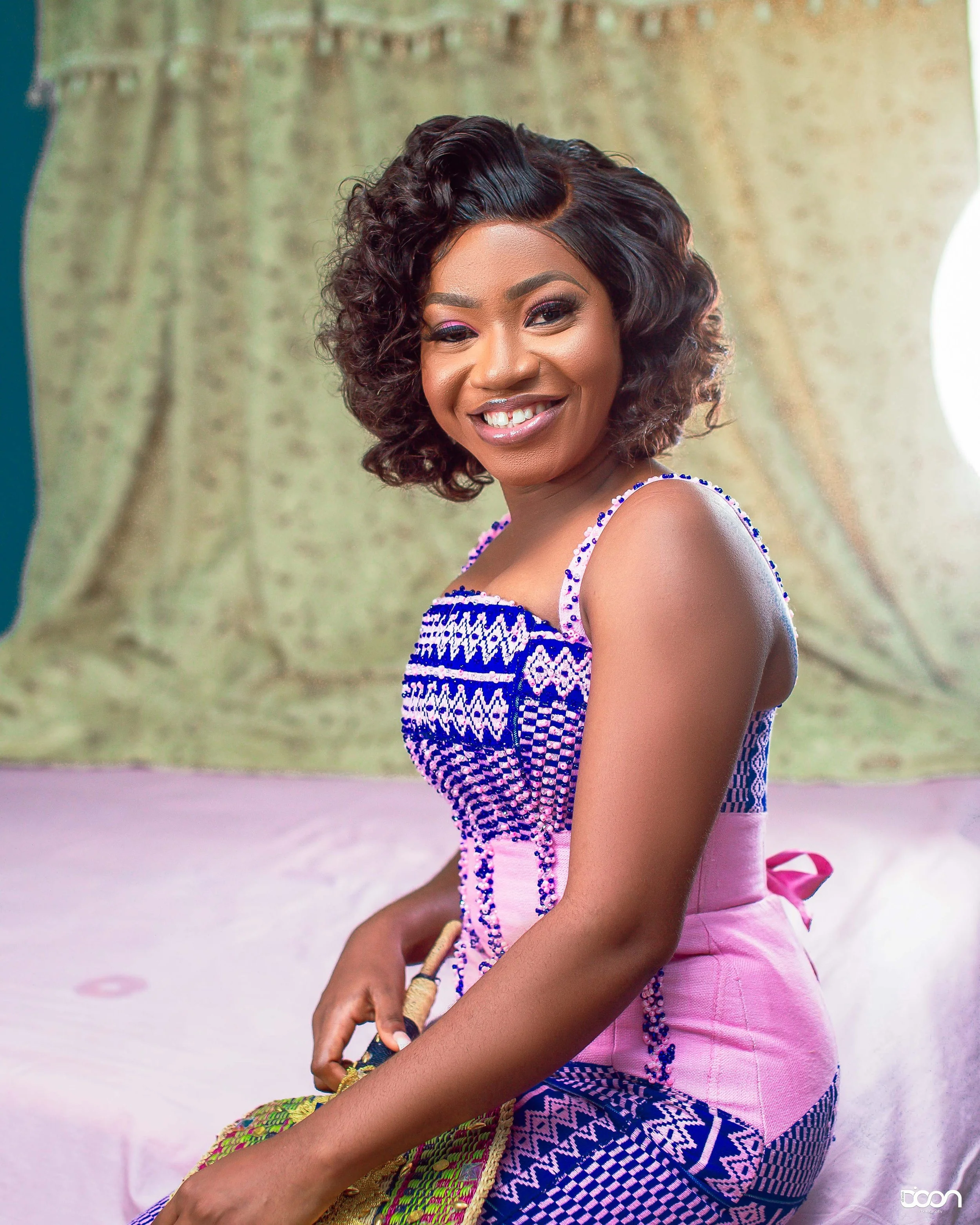 A woman in a colorful, patterned dress smiling while sitting on a bed with a pink bedspread, against a backdrop of green curtains and a bright light source.