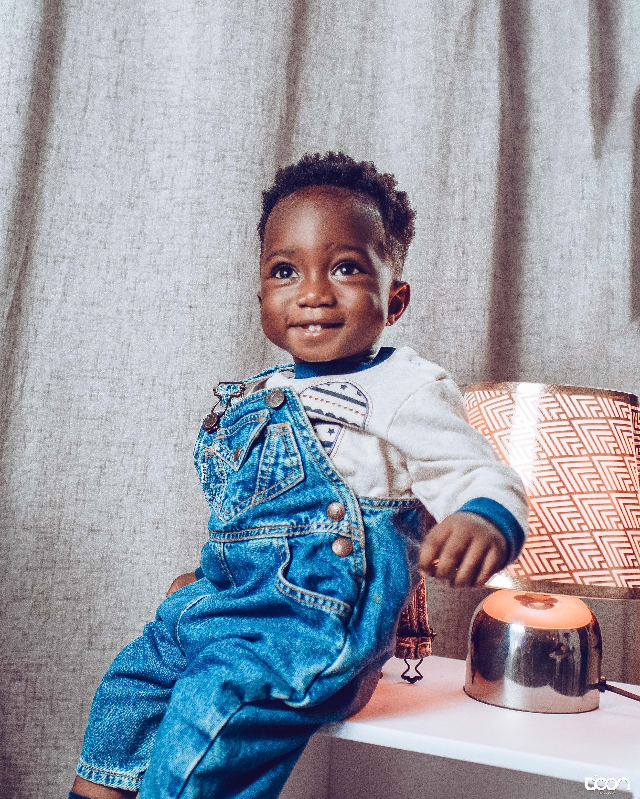 Smiling young boy sitting on a bedside table near a modern lamp with a patterned lampshade, wearing denim overalls and a long-sleeve shirt, in front of a beige curtain.