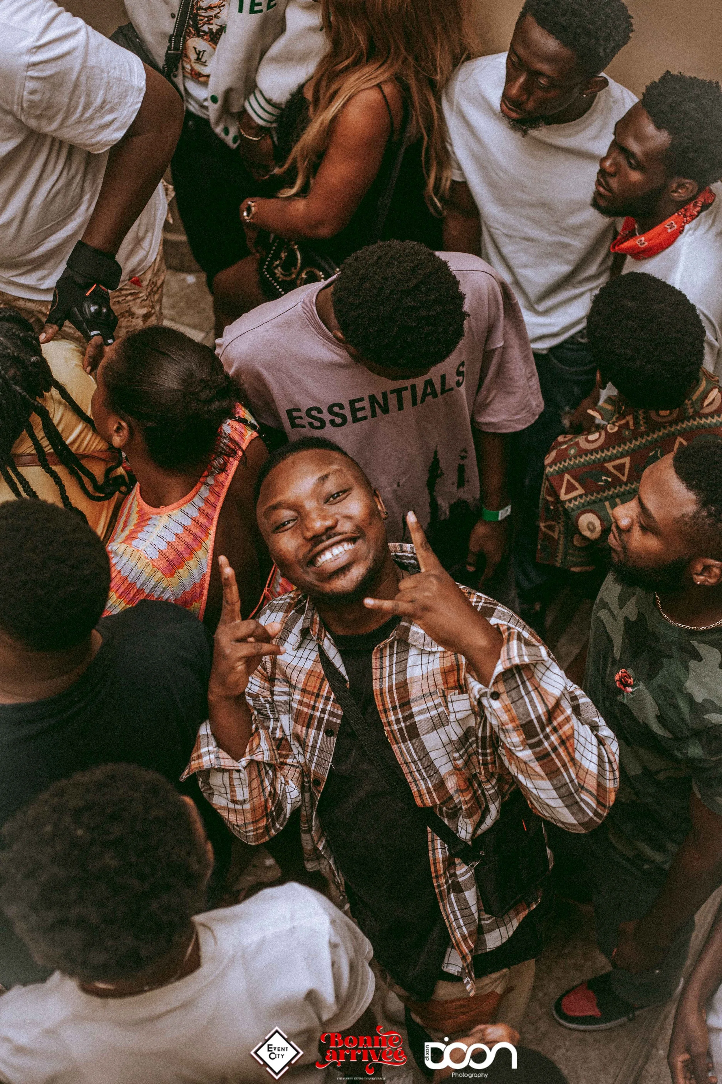 A group of young people, mostly men, gathered in a crowded setting, with one young man in the front smiling and showing a hand gesture, surrounded by other people.
