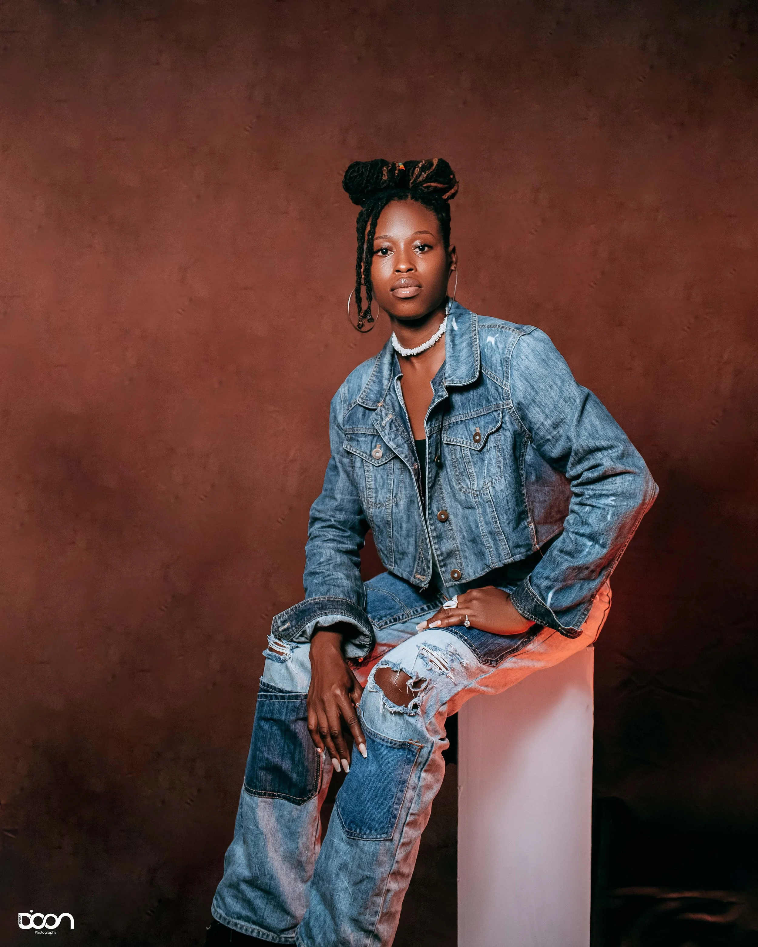 A woman sitting on a white block in front of a brown textured background, wearing a blue denim jacket and ripped jeans, with her hair styled in twists, accessorized with hoop earrings and a choker necklace.
