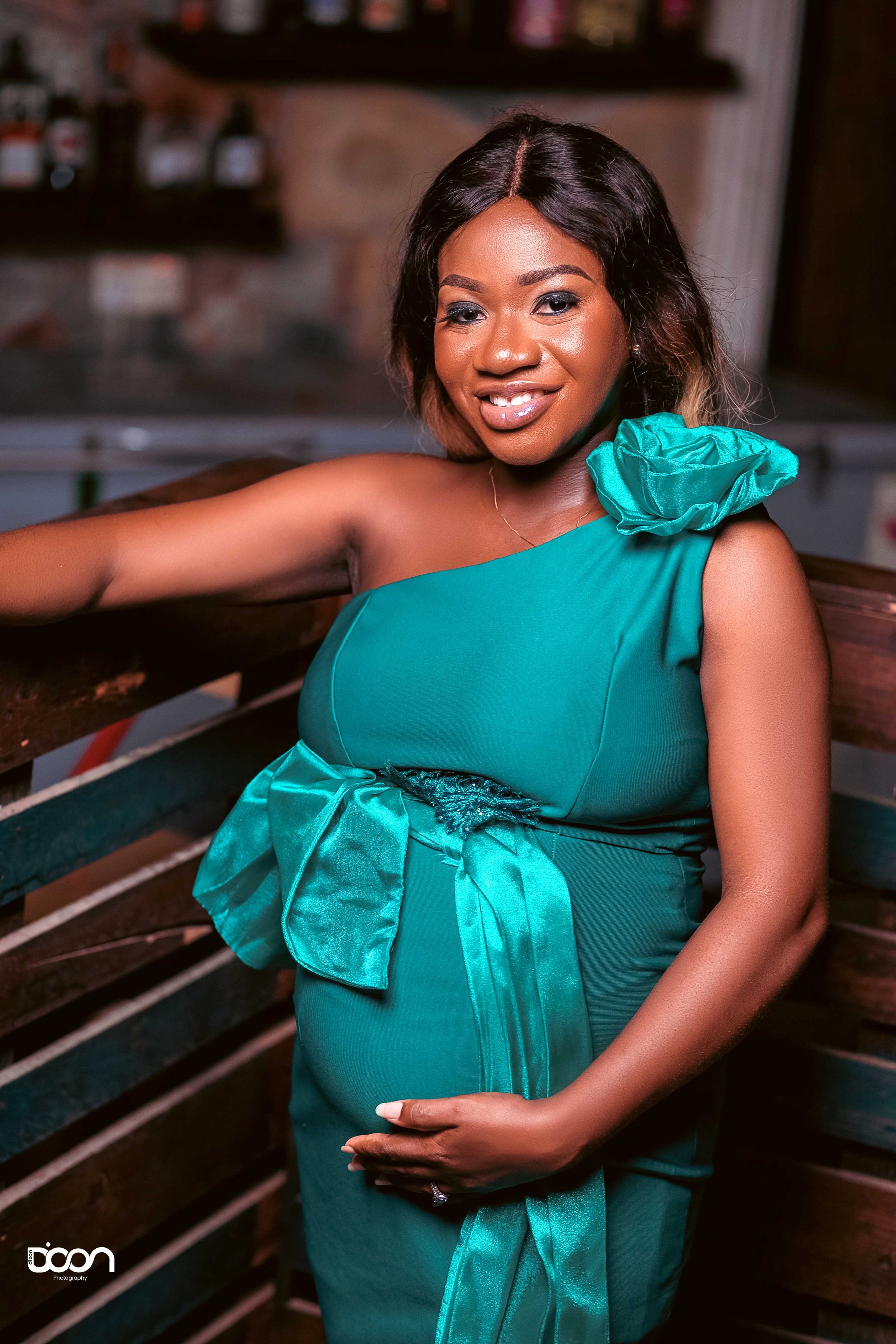 A woman in a teal dress with a ruffled shoulder and a large bow, smiling and gently resting her hand on her pregnant belly, posing indoors.
