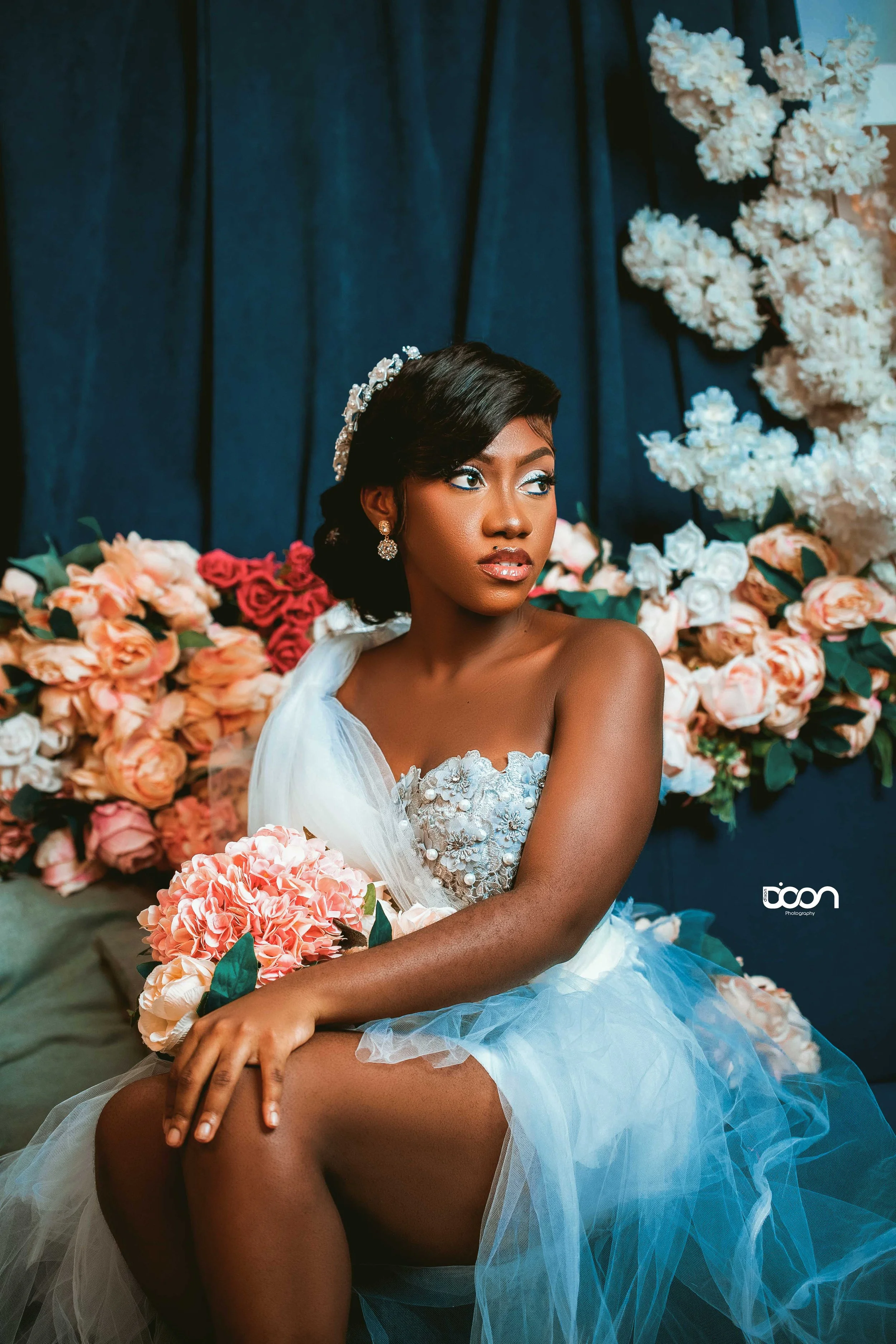 Young woman in wedding dress sitting amidst pink and white roses, holding a bouquet, with dark curtain backdrop and white floral decoration.
