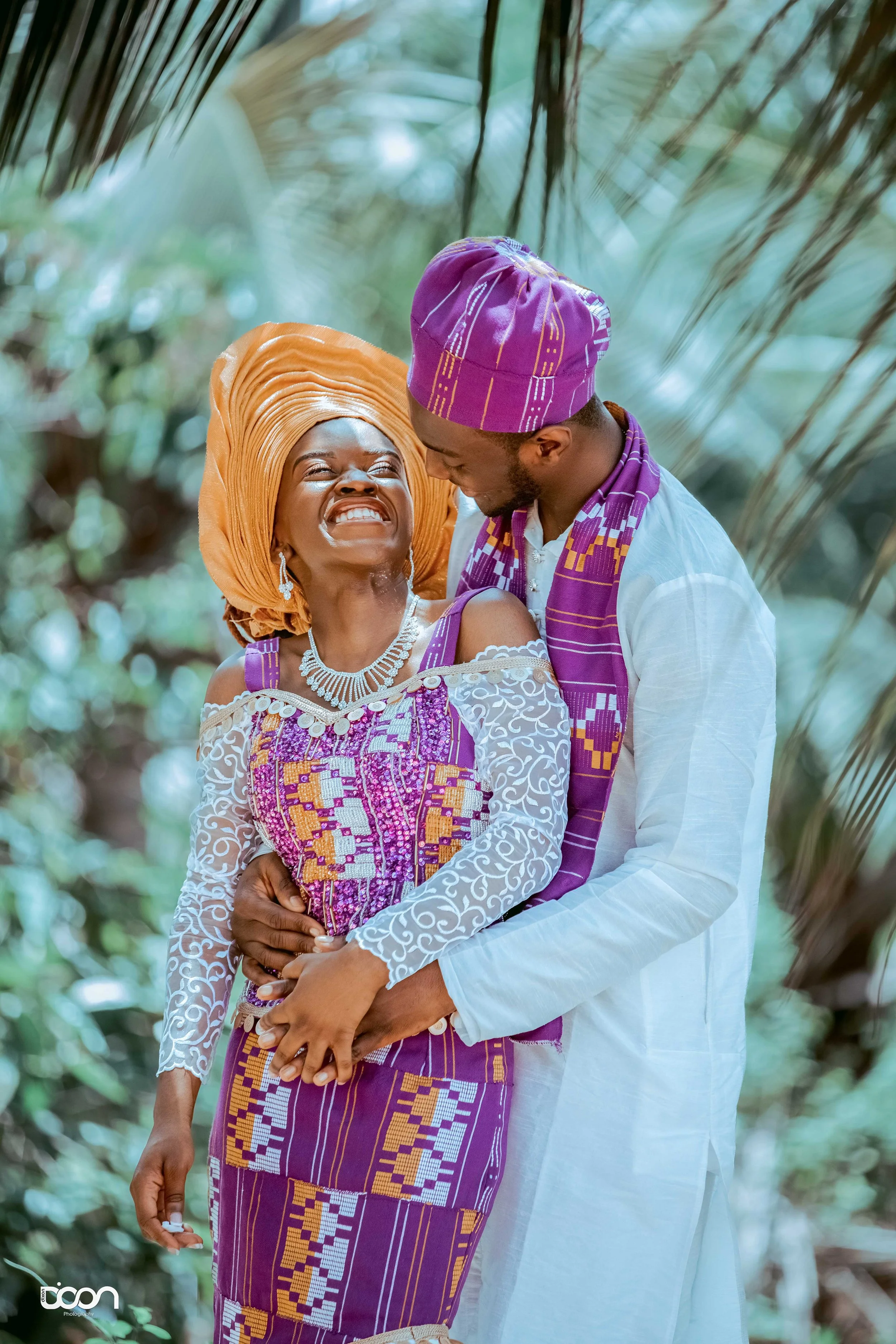 A joyful couple dressed in traditional West African attire, sharing a loving moment outdoors surrounded by greenery and palm leaves.