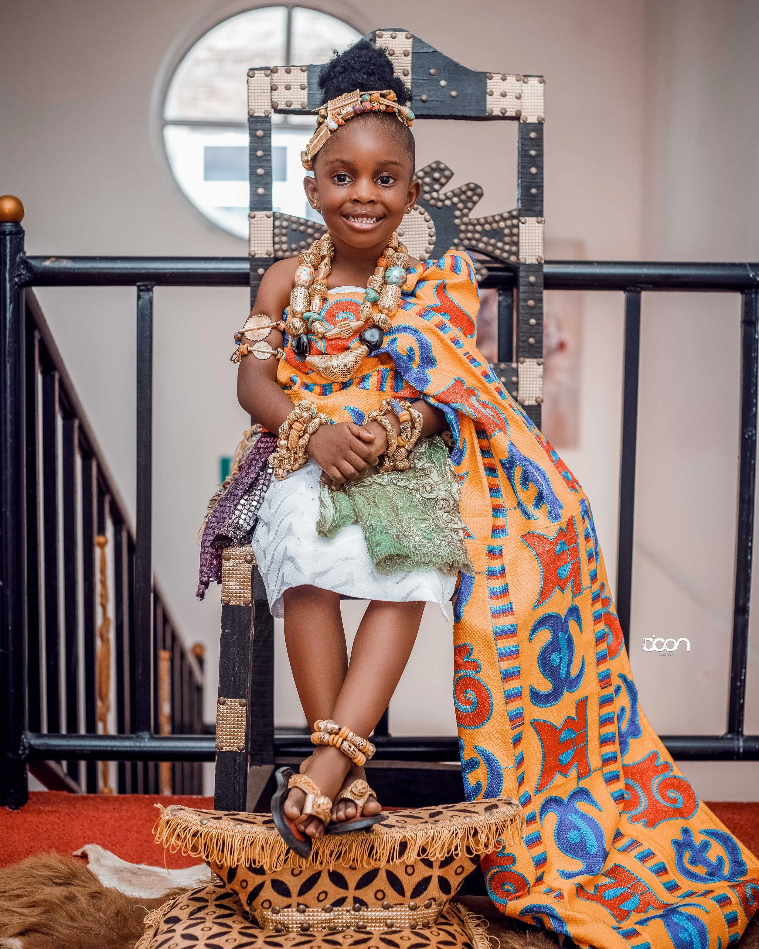 Young girl dressed in traditional African attire adorned with jewelry, sitting on a platform, smiling at the camera inside a room with a round window in the background.