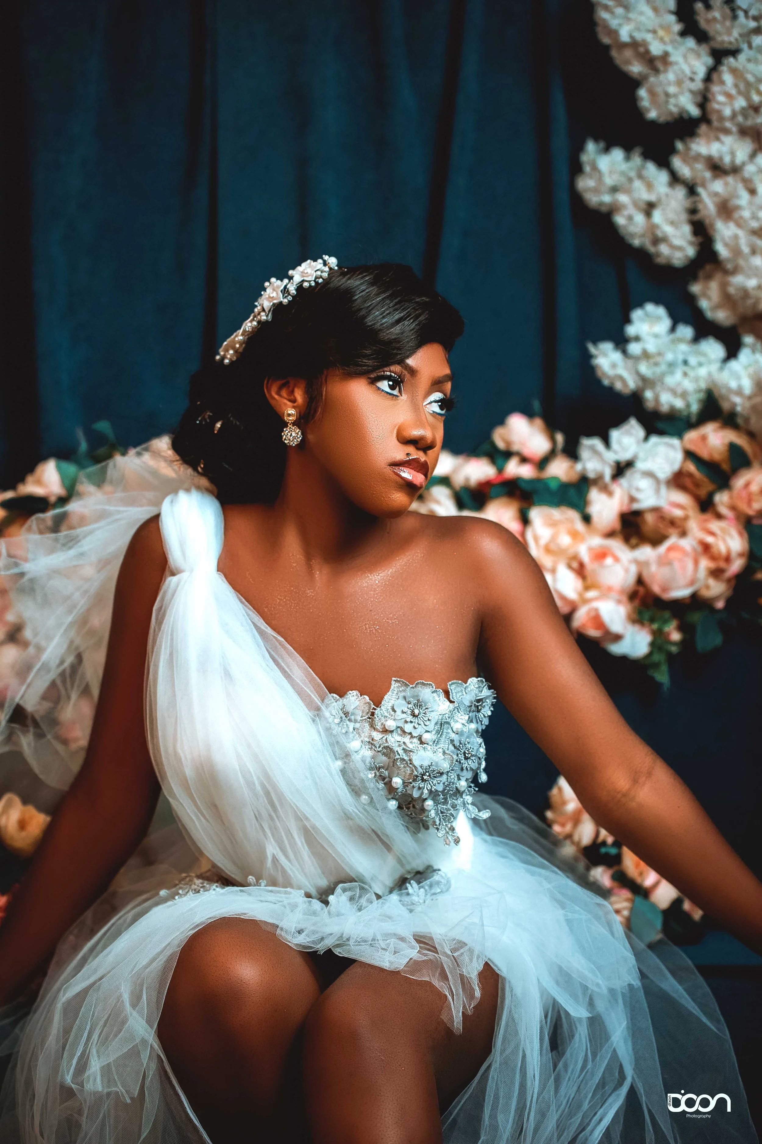 A woman dressed in a white, embellished dress with tulle fabric, sitting in front of a backdrop of dark blue curtains and pink and white flowers, wearing earrings and a jeweled headpiece.