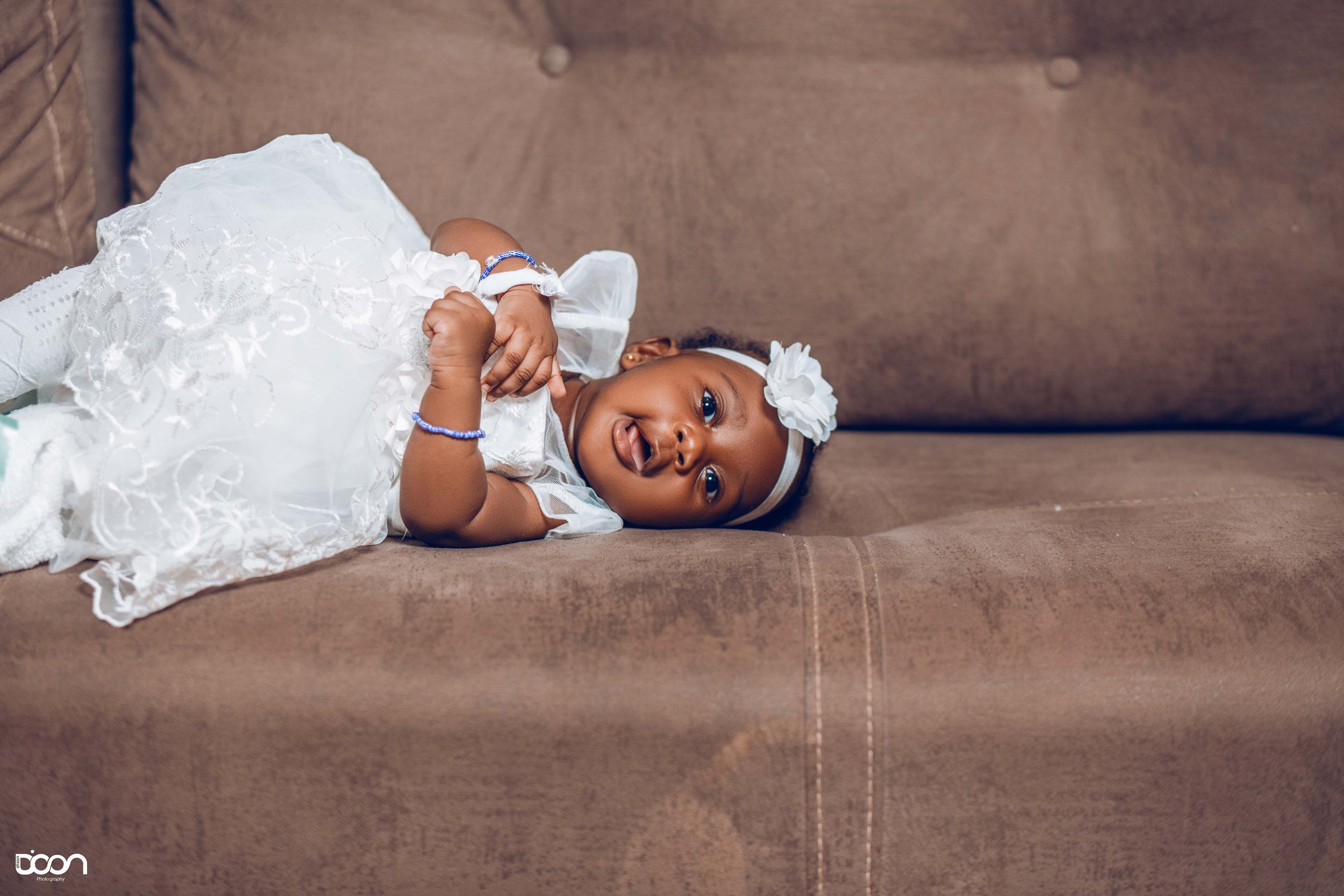 A young girl lying on a brown couch, wearing a white dress with lace details and a white flower headband, smiling and looking at the camera.