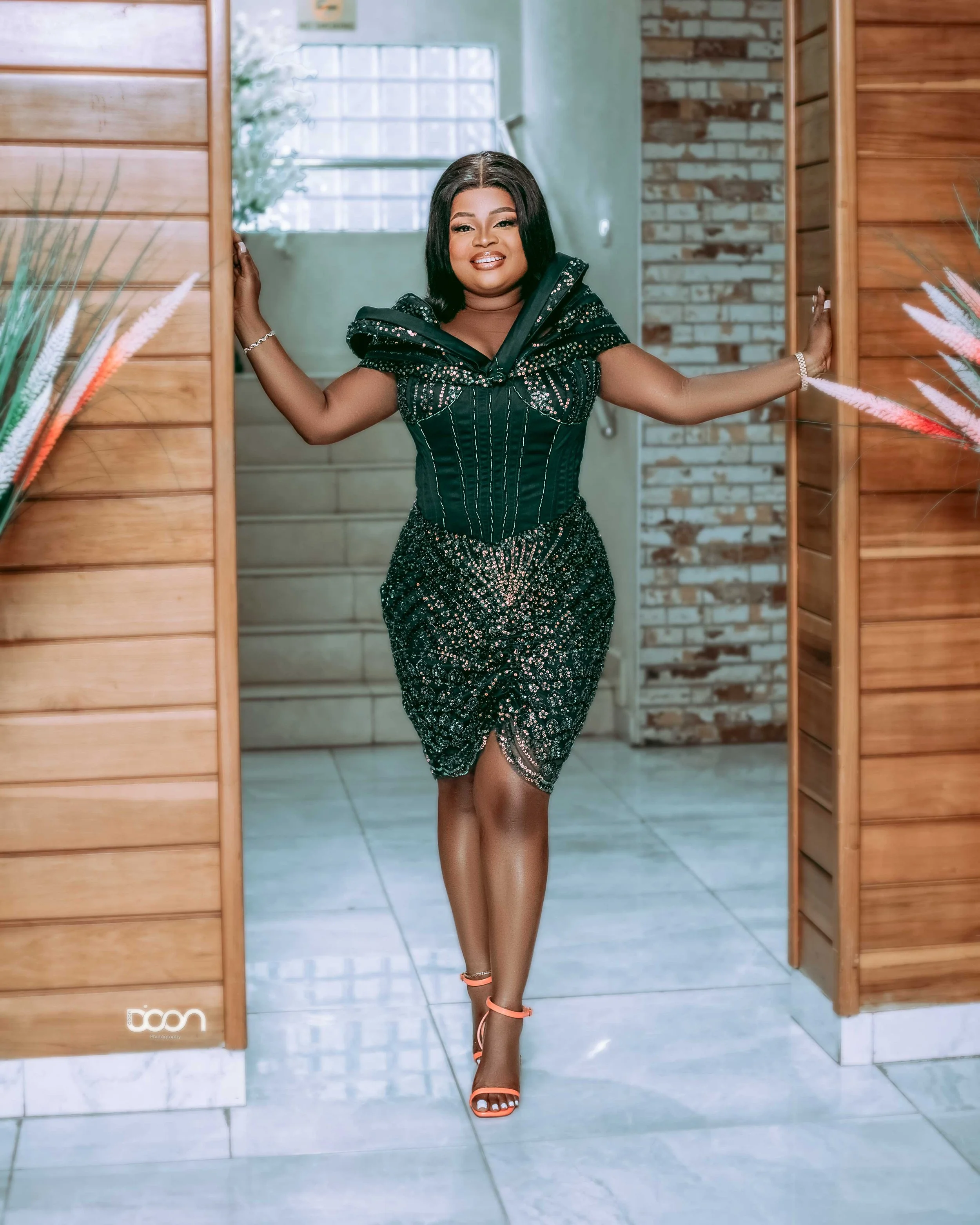 A woman in a black, embellished dress stands with arms outstretched in a doorway, smiling at the camera.