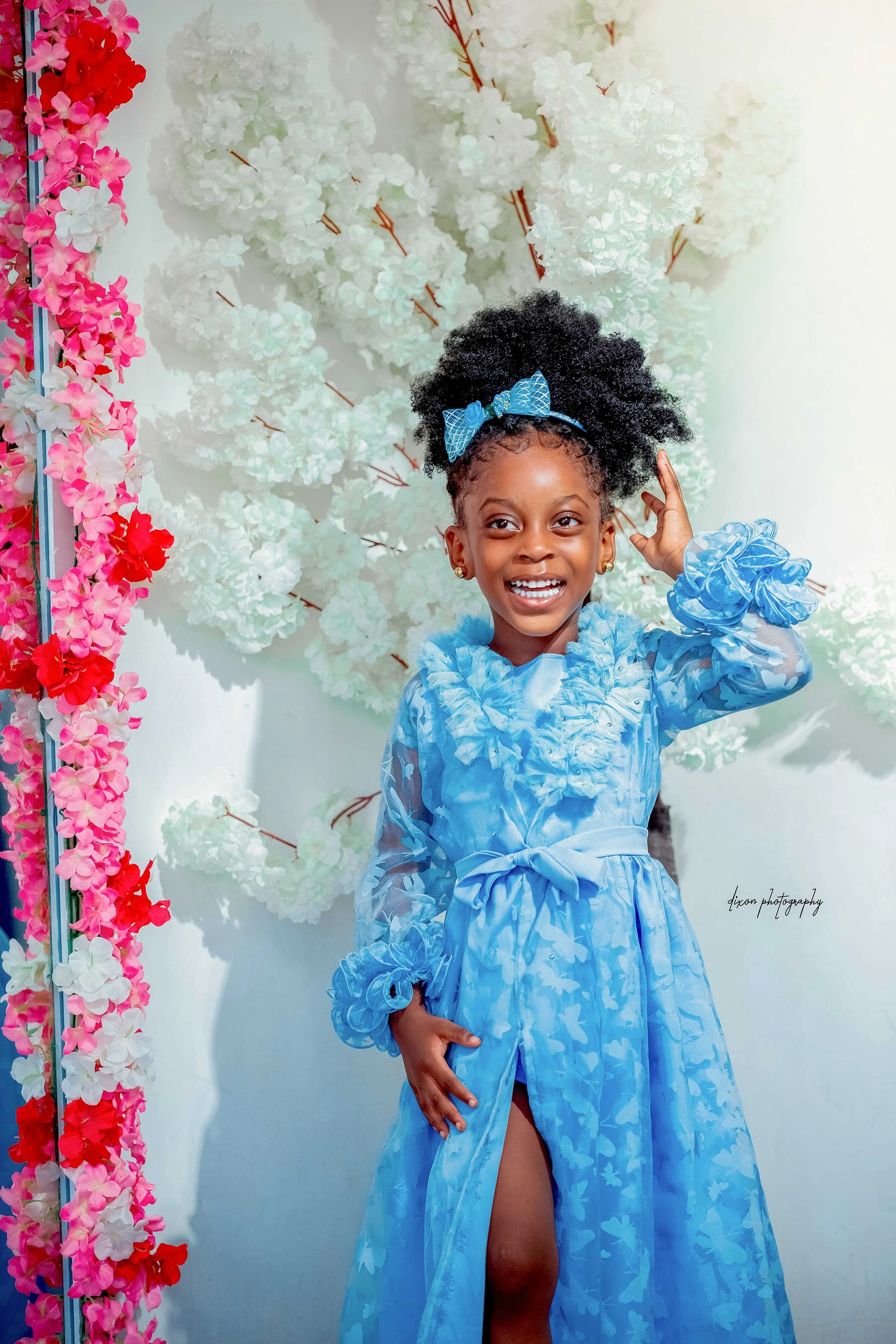 A young girl smiling, wearing a light blue dress with ruffles and a bow, standing in front of a wall decorated with white and pink flowers. She has a blue bow headband and gold earrings.