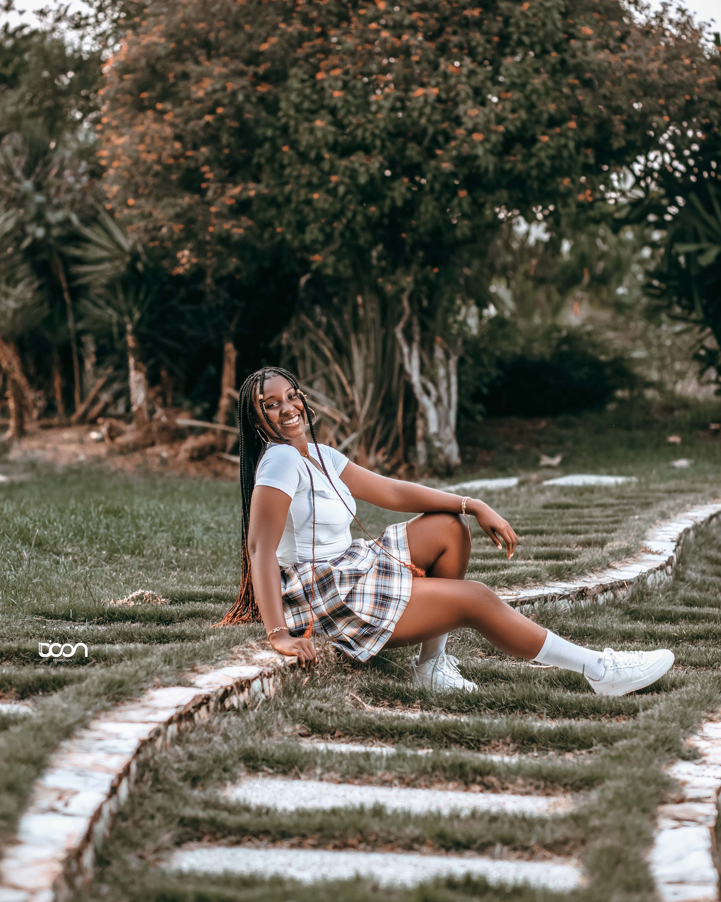 Smiling young woman with braided hair sitting on a garden path, wearing a white t-shirt, plaid skirt, white sneakers, and knee-high socks, with trees and bushes in the background during late afternoon.