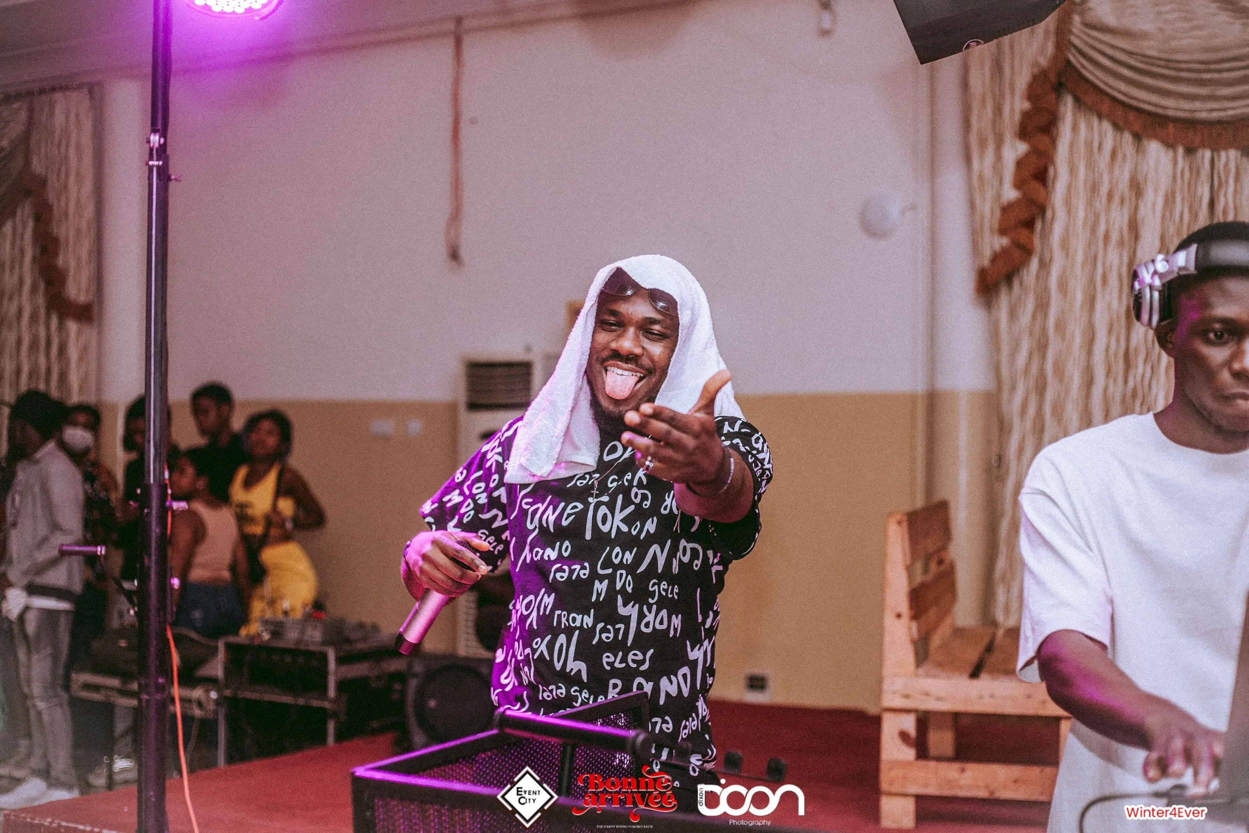 Man with a towel on his head, sticking out his tongue and smiling, holding a microphone at an indoor event.
