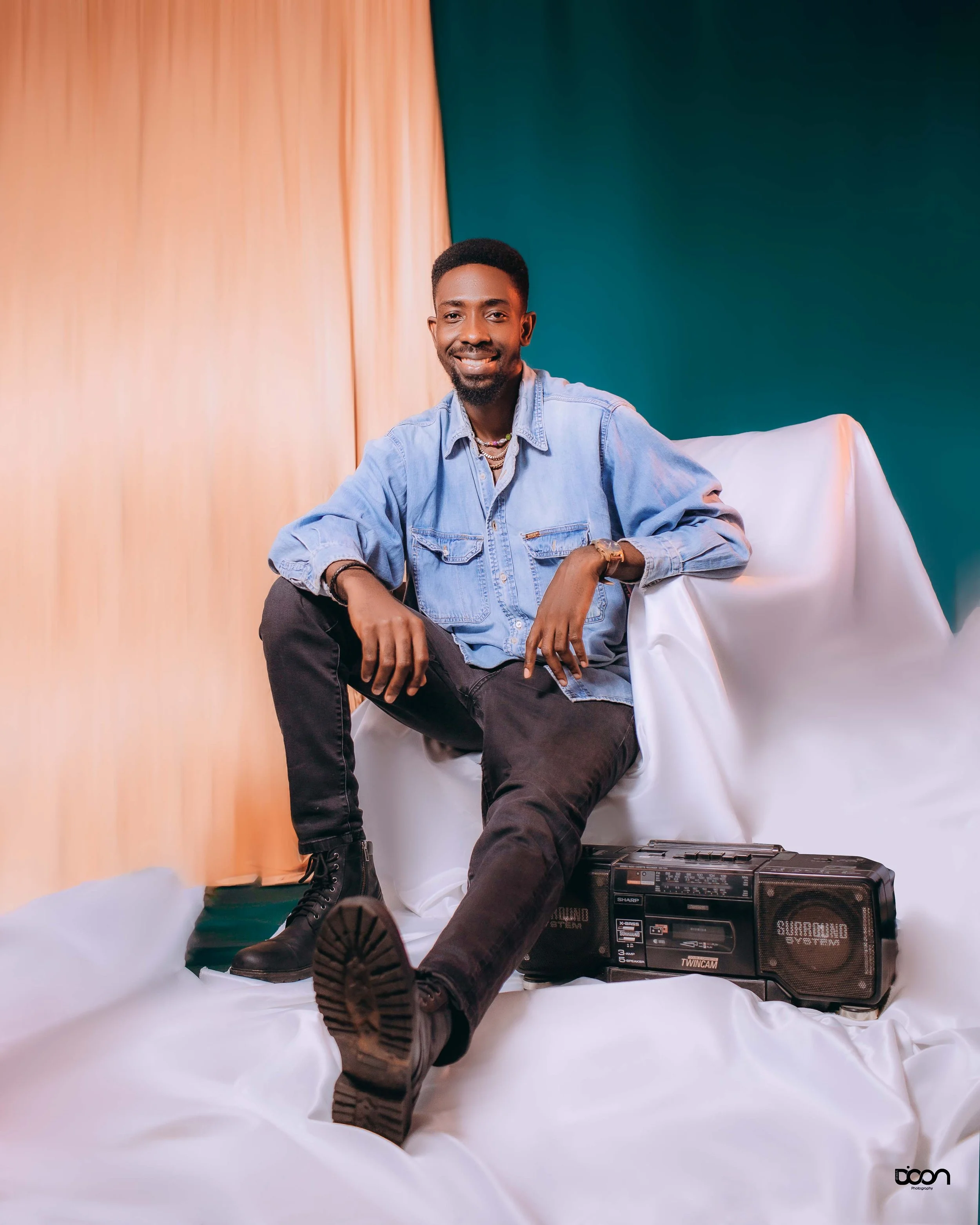 A young man sitting on a white draped surface with one leg extended, wearing black boots, black pants, a light blue denim shirt, accessorized with layered necklaces and a watch, smiling at the camera, with a vintage boombox placed beside him, against