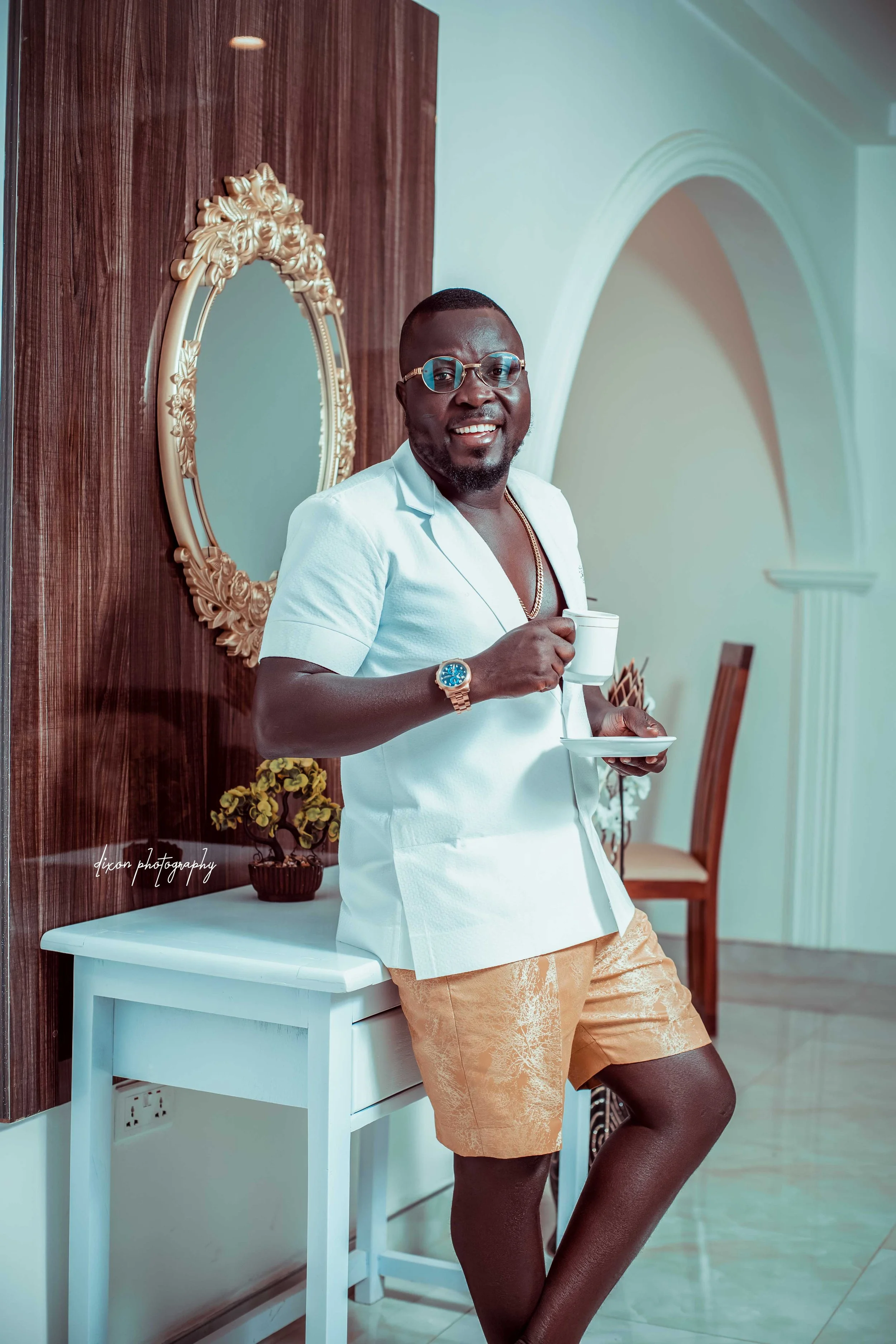 A smiling man in a white shirt, gold shorts, and glasses standing indoors, holding a cup and saucer.