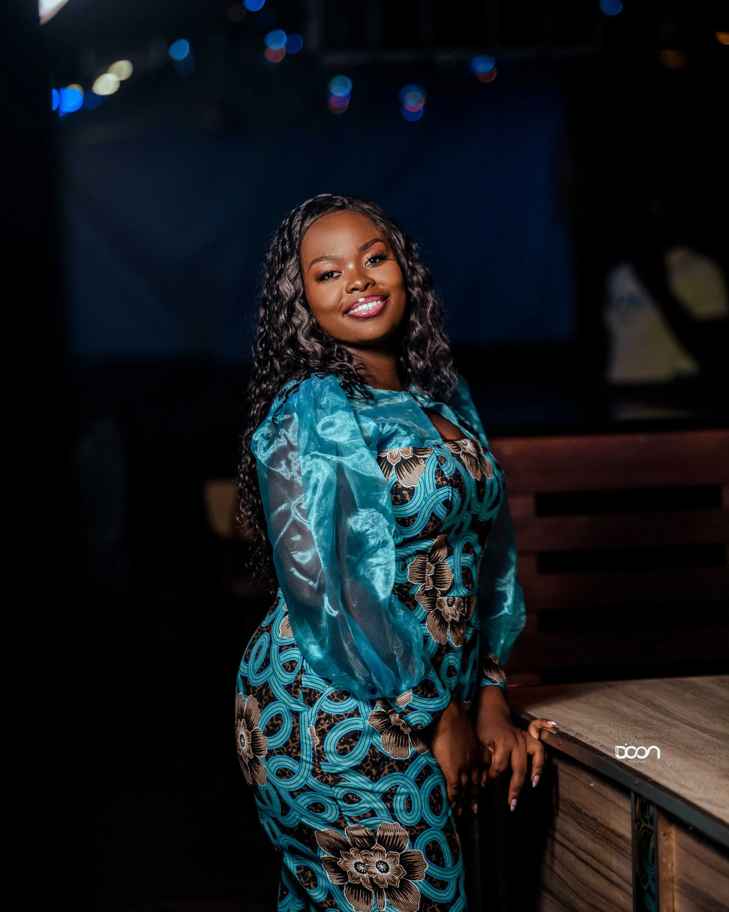 A woman with long curly black hair smiling, wearing a blue patterned dress and a matching sheer blue jacket, standing next to a wooden table in a dark room with a blurred background.
