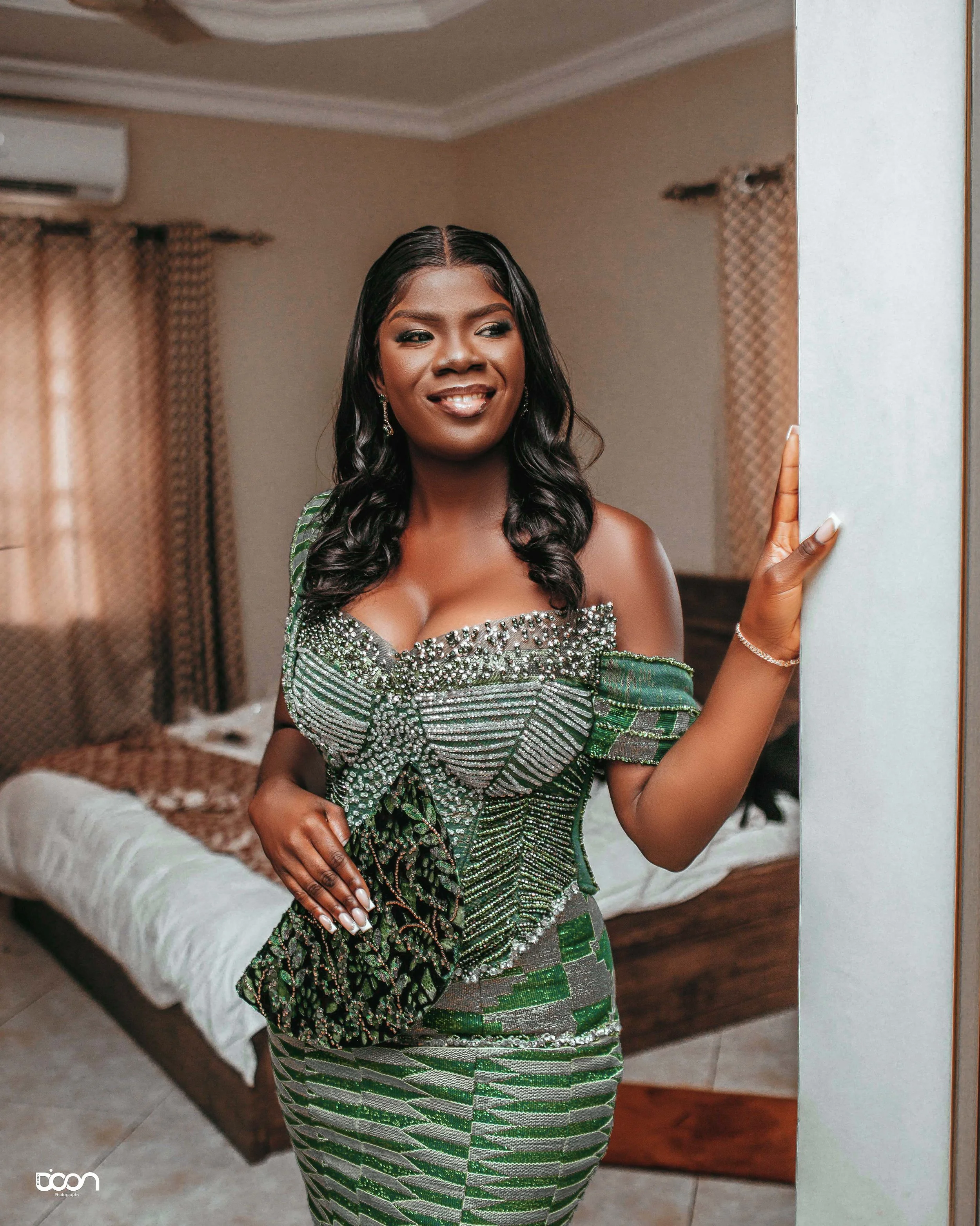 A woman dressed in an ornate green and silver traditional outfit standing in a bedroom, smiling and looking away from the camera.