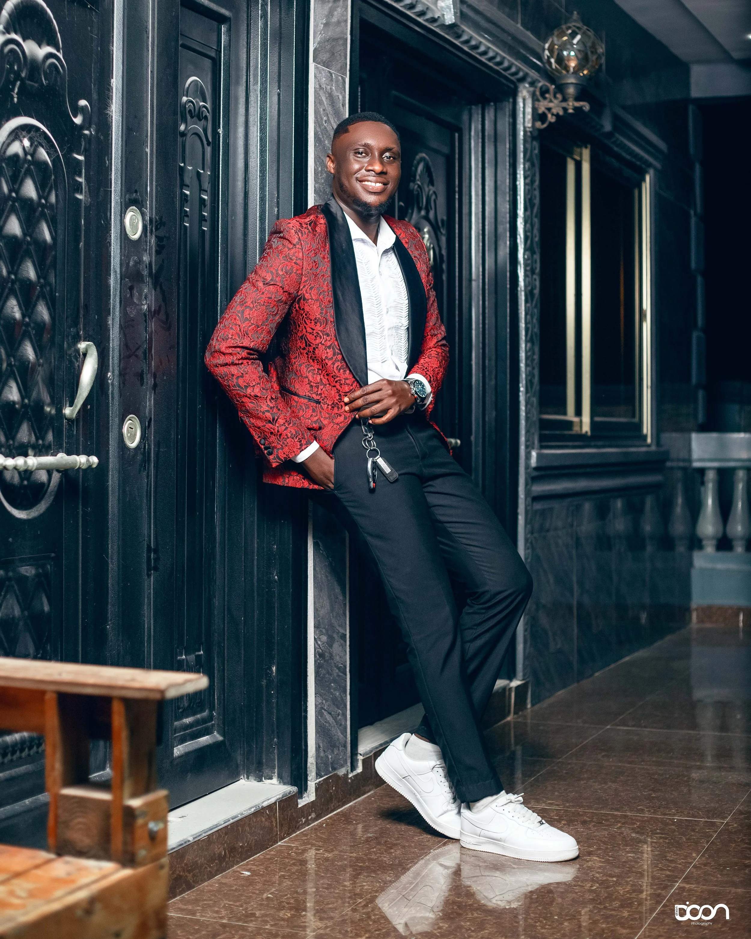 A man in a red patterned blazer, white shirt, black pants, and white sneakers leaning against a black wall in a stylish indoor setting, smiling at the camera.