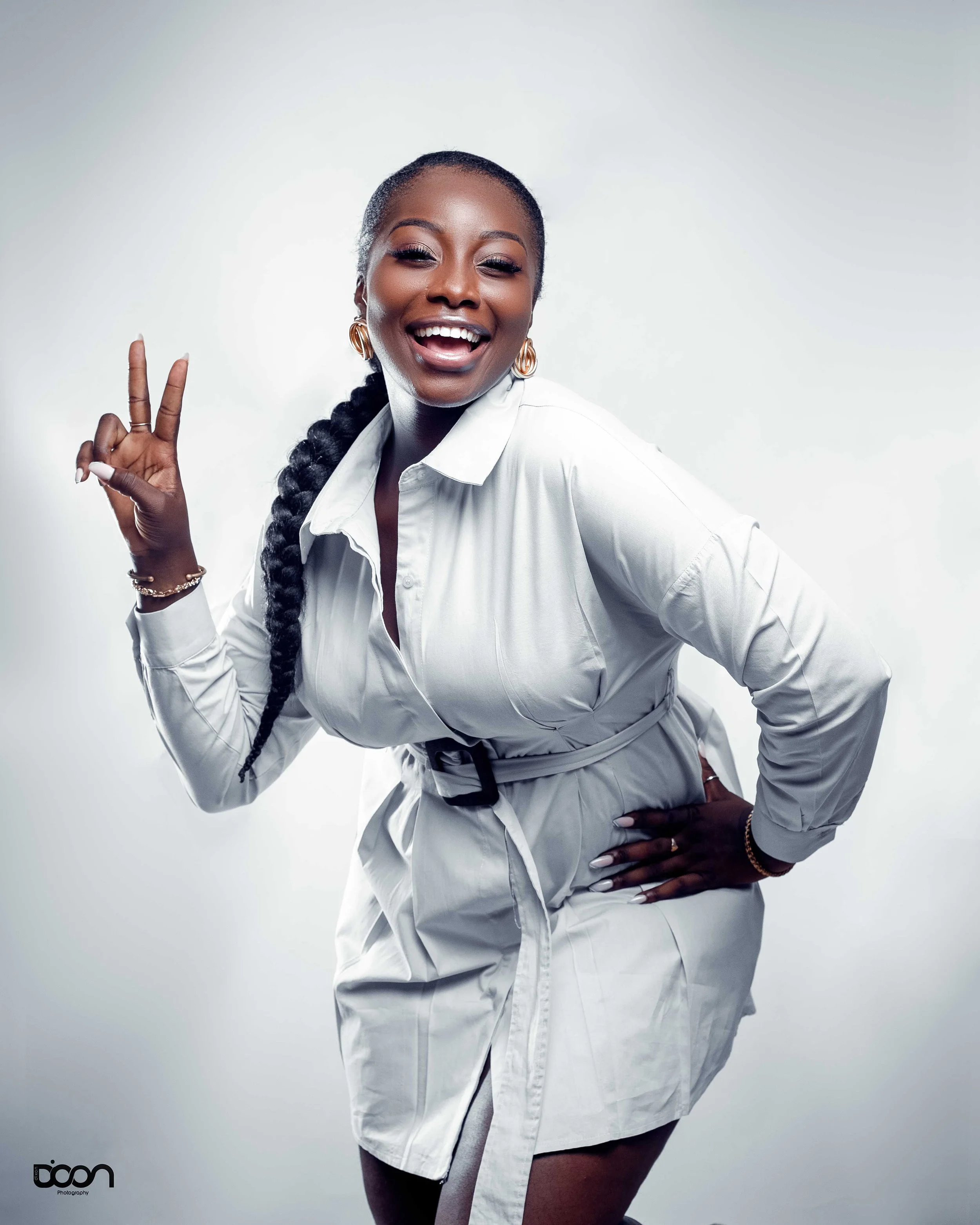 Woman smiling and making a peace sign with her right hand, wearing a white long-sleeve dress with a buckle belt, earrings, and bracelets, against a plain light background.