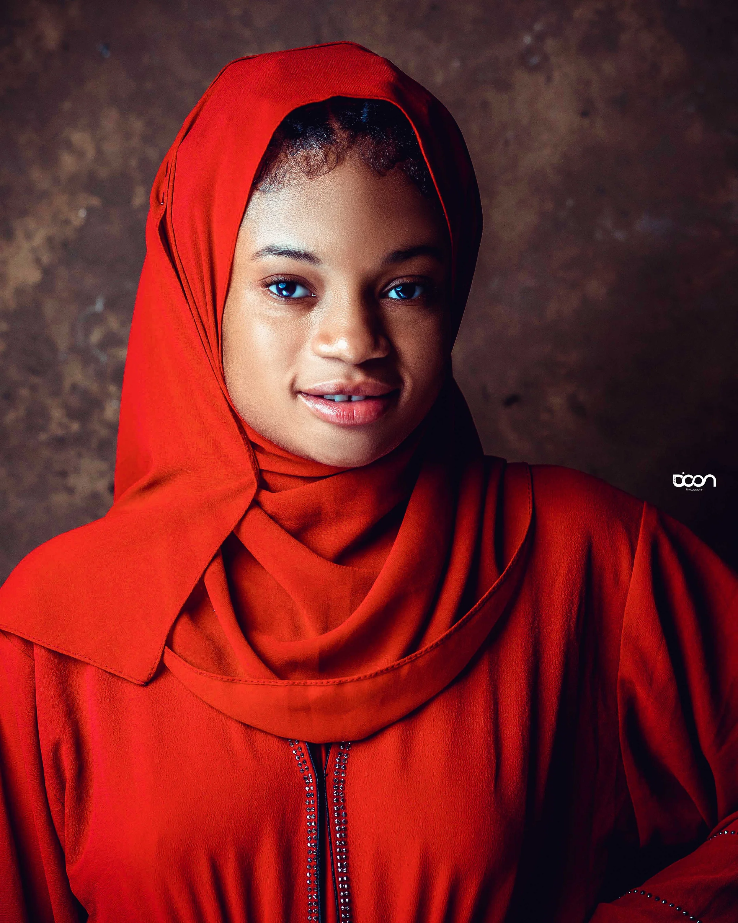 A woman with dark curly hair, bright blue eyes, and a warm smile, wearing a red hijab and matching red jacket, standing against a textured brown background.