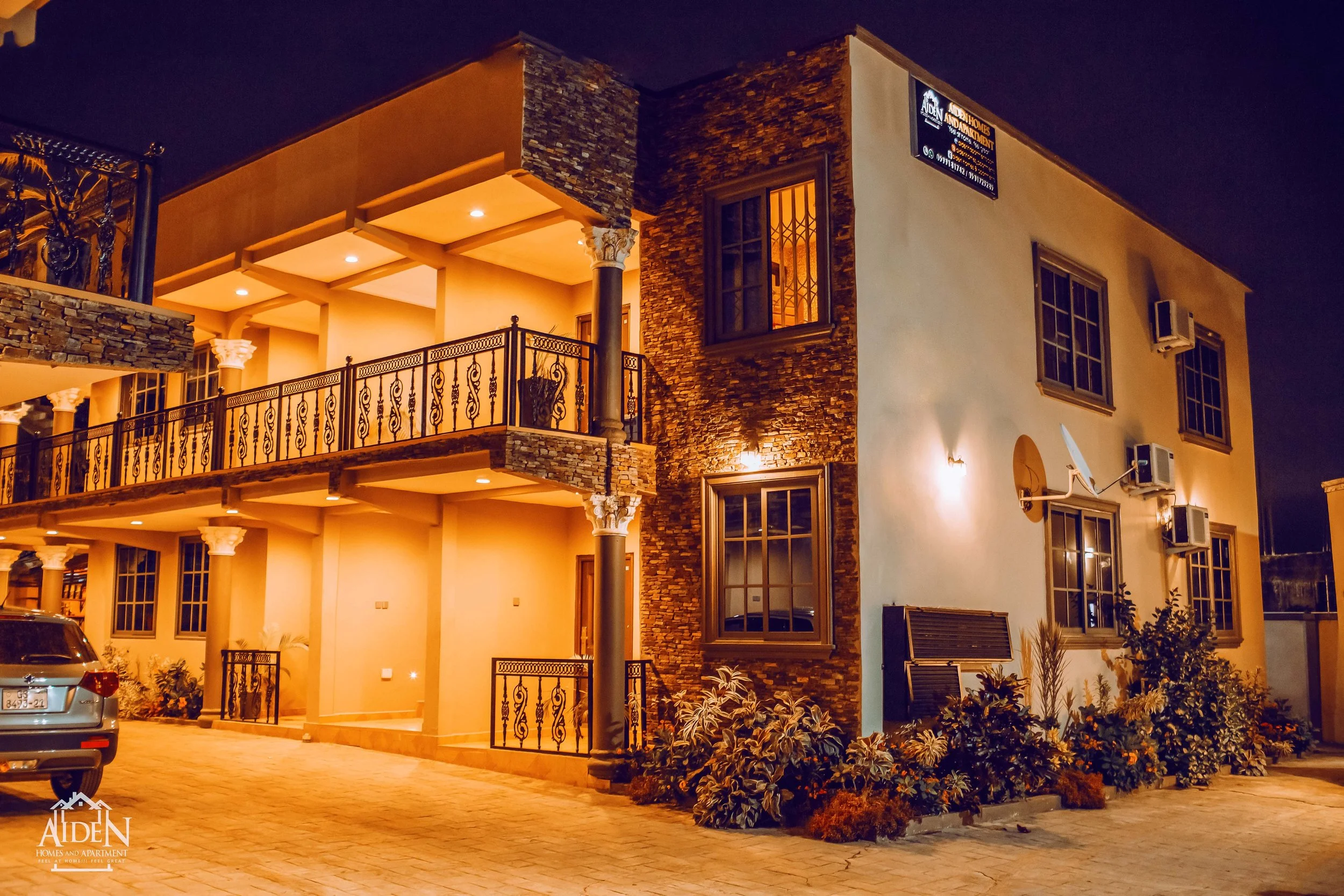 Exterior of a multi-story apartment building at night with illuminated windows, balcony railings, and landscaped bushes in front.