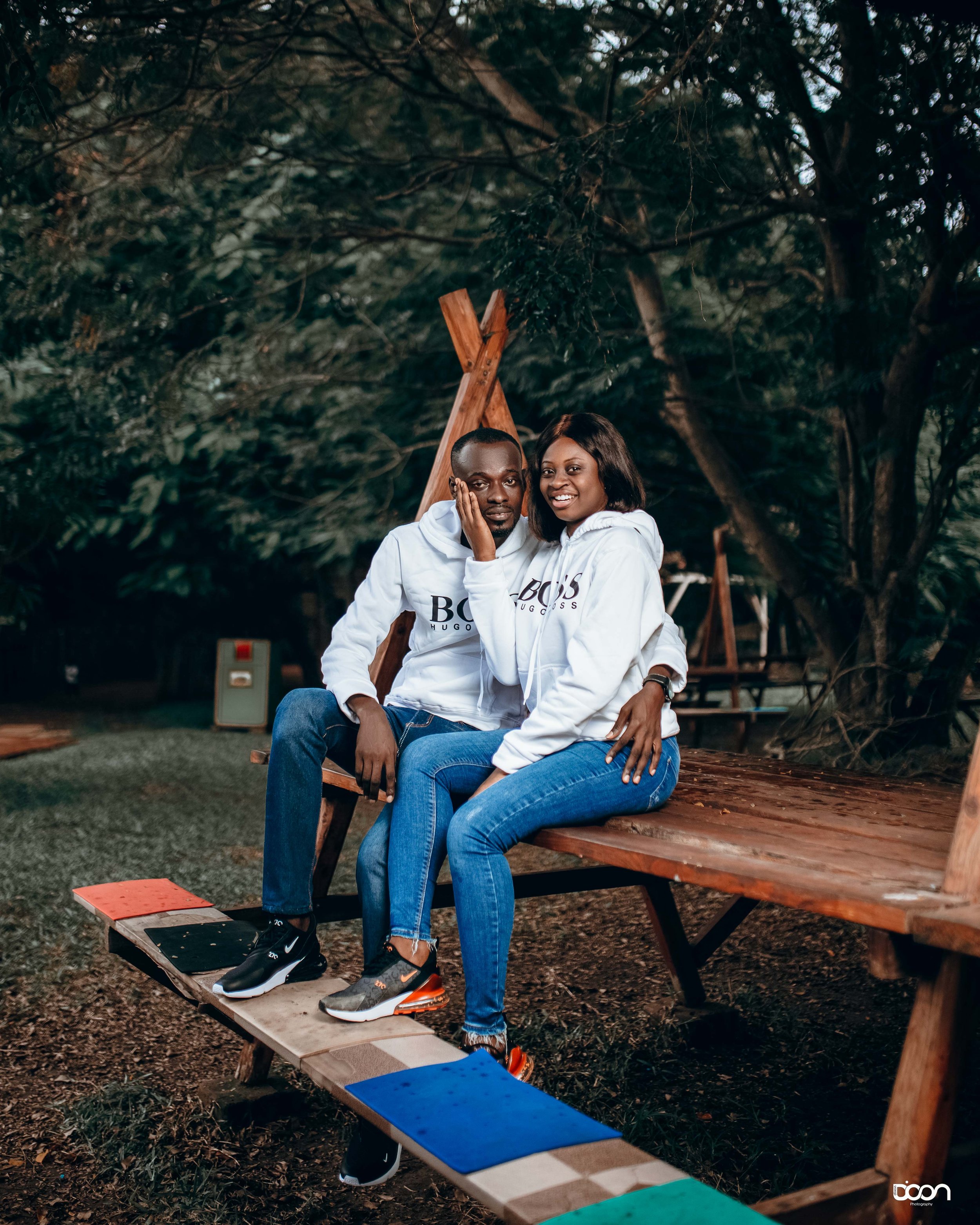 A man and woman sit closely on a wooden bench in a park, wearing matching white hoodies with 'BOSS' logo, surrounded by trees, in an outdoor setting at dusk.