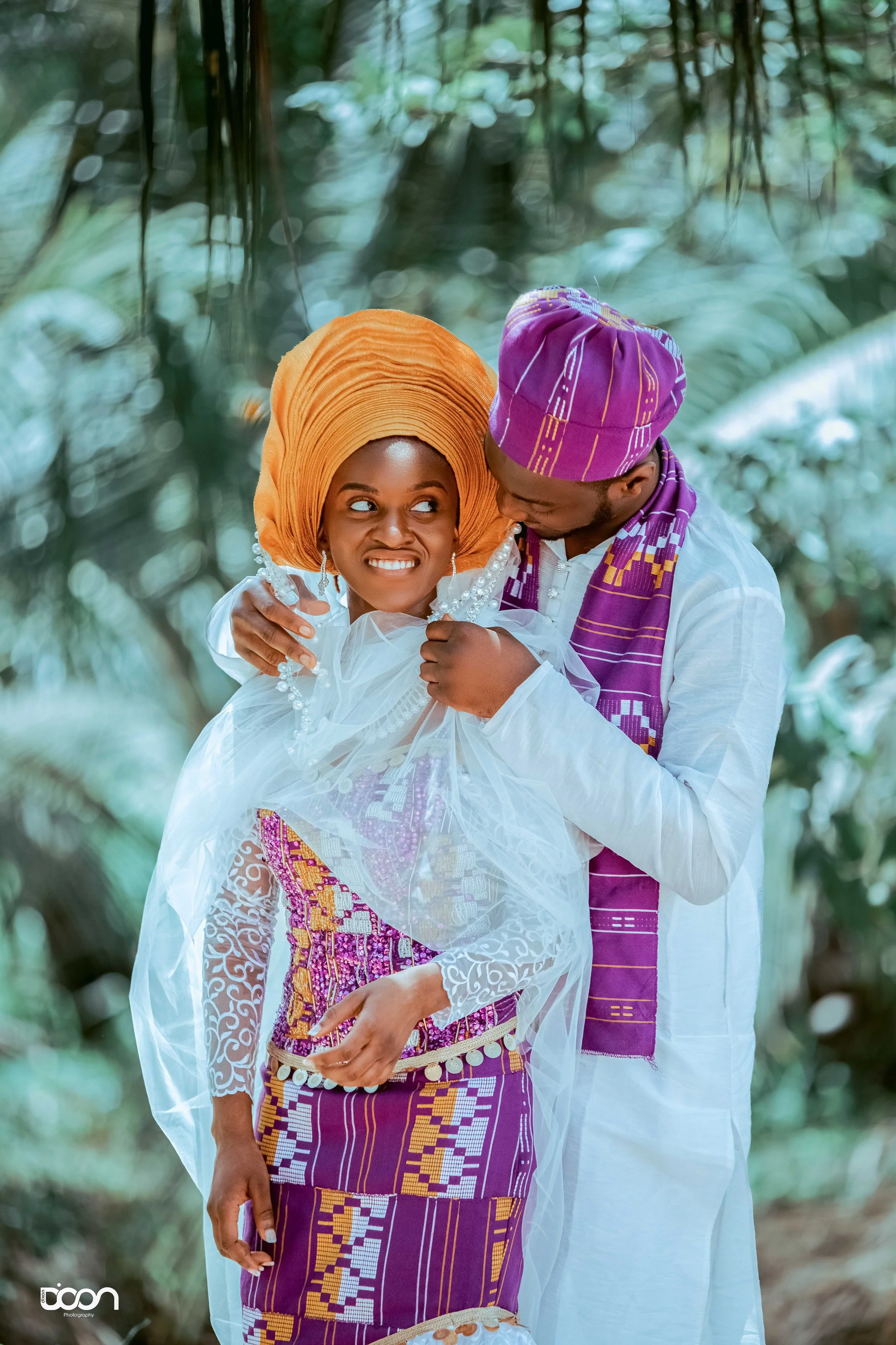 A couple dressed in traditional African attire, the woman in a purple and white patterned dress with a matching headwrap, and the man in a white outfit with a purple and gold headwrap, share an intimate moment amidst a lush green forested environment