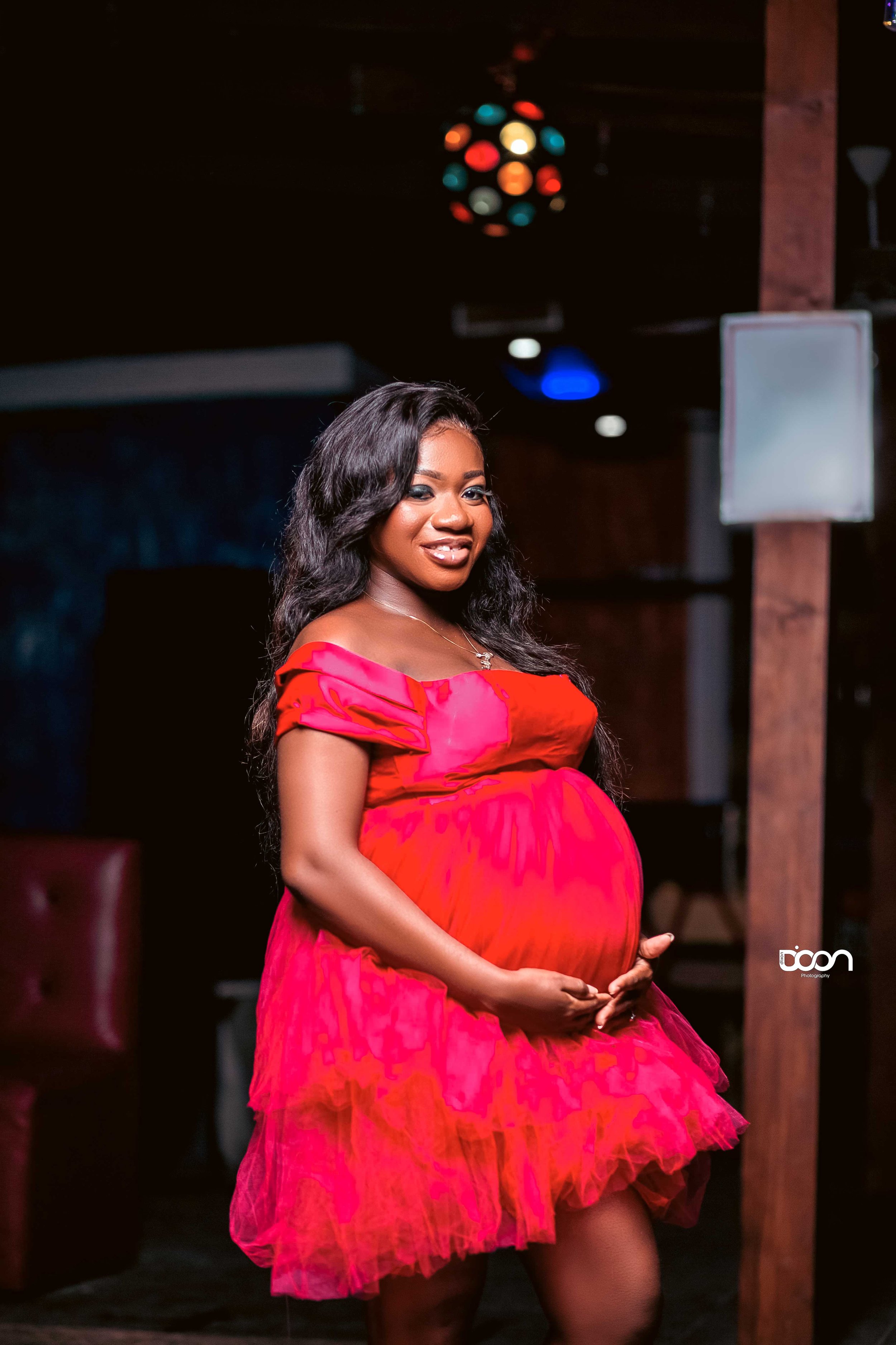 A pregnant woman wearing a bright pink dress, smiling and holding her belly, standing indoors with colorful lights overhead.