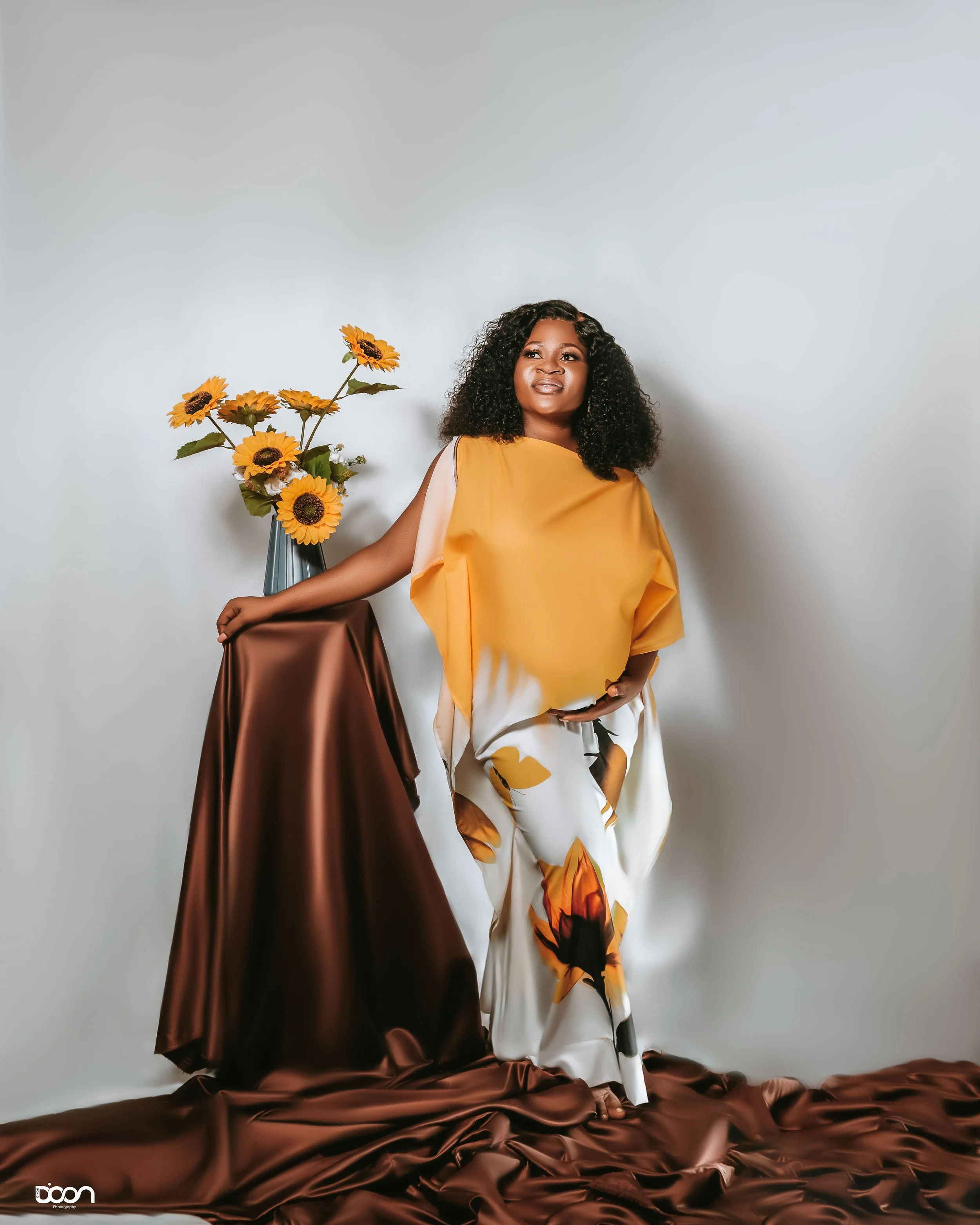A woman with curly hair wearing a yellow top and floral pants stands beside a table covered with brown satin fabric, with a sunflower flower arrangement on the table, against a plain light background.