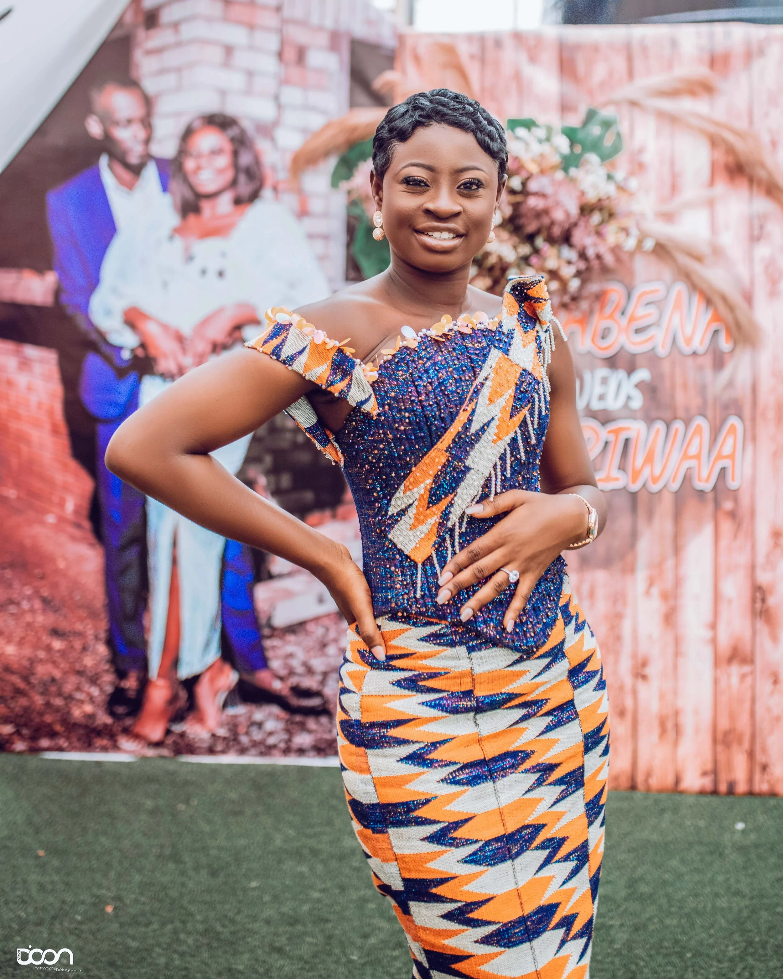 A woman in a colorful, patterned dress facing the camera, standing with one hand on her hip in front of a decorated backdrop with a photo of a couple and text.