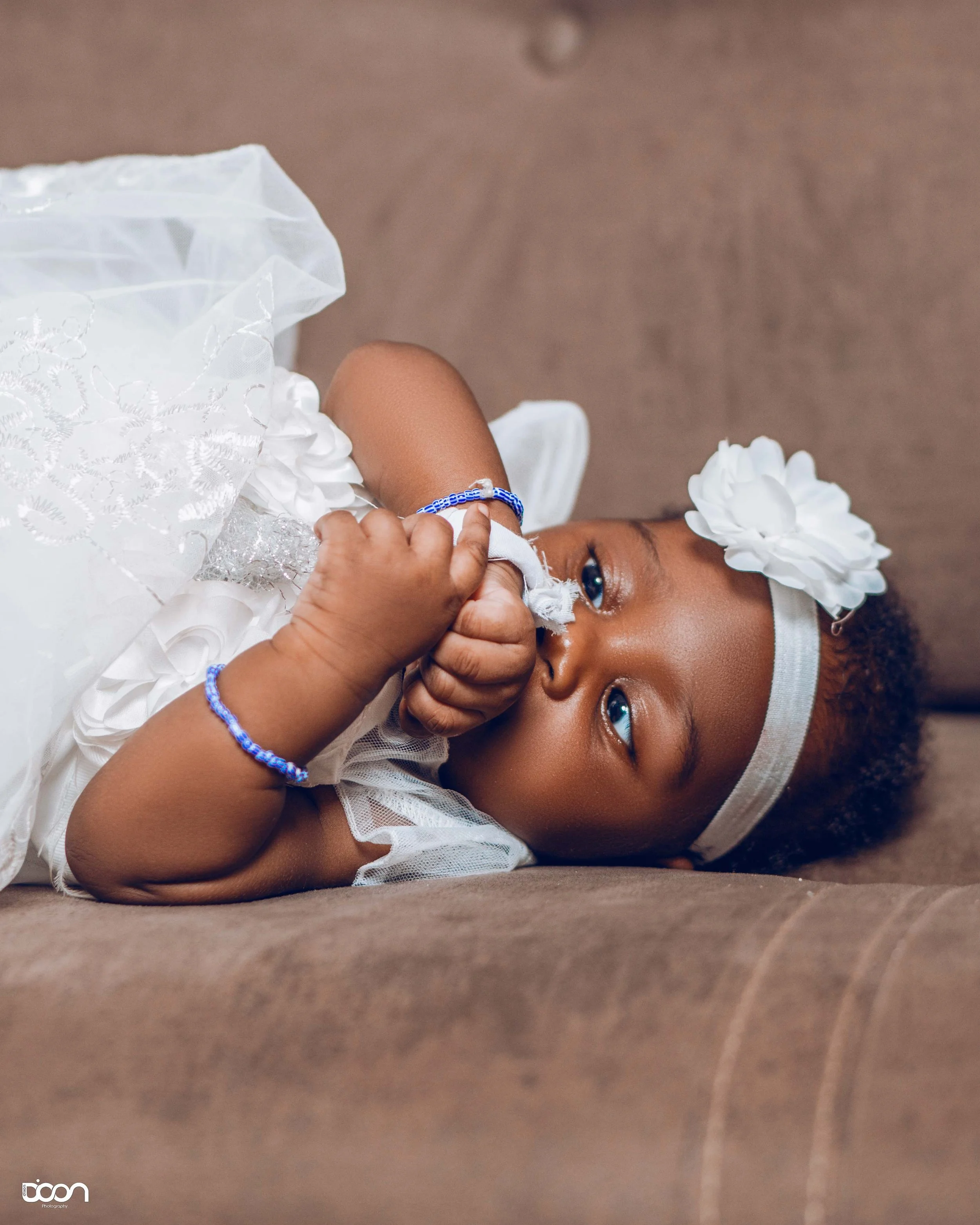 A young girl lying on a brown surface, wearing a white dress and a white headband with a large white flower, holding her hand near her face, with a calm expression and bright blue eyes.