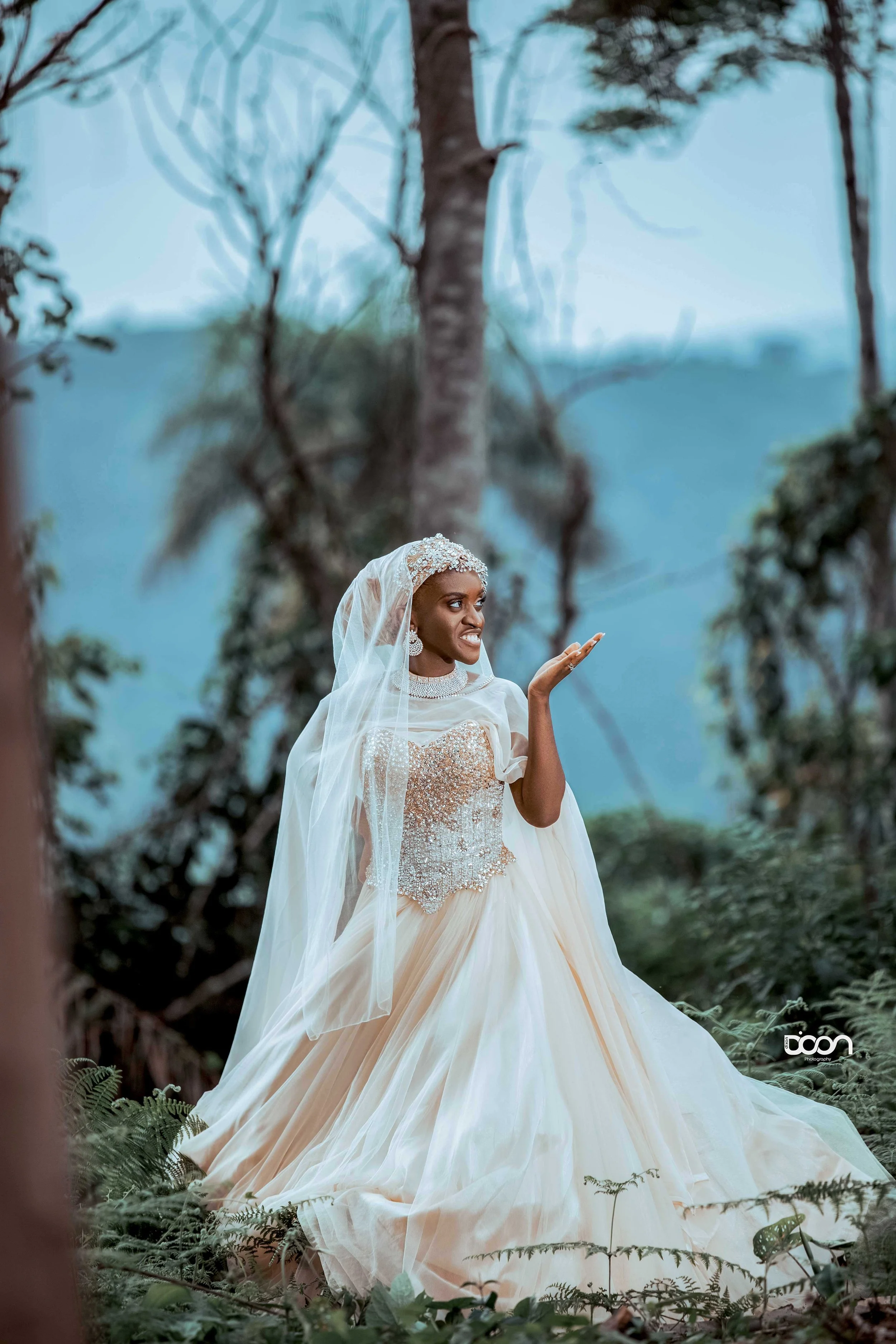 A woman dressed in a wedding gown with a veil, standing outdoors in a forest with trees and ferns.