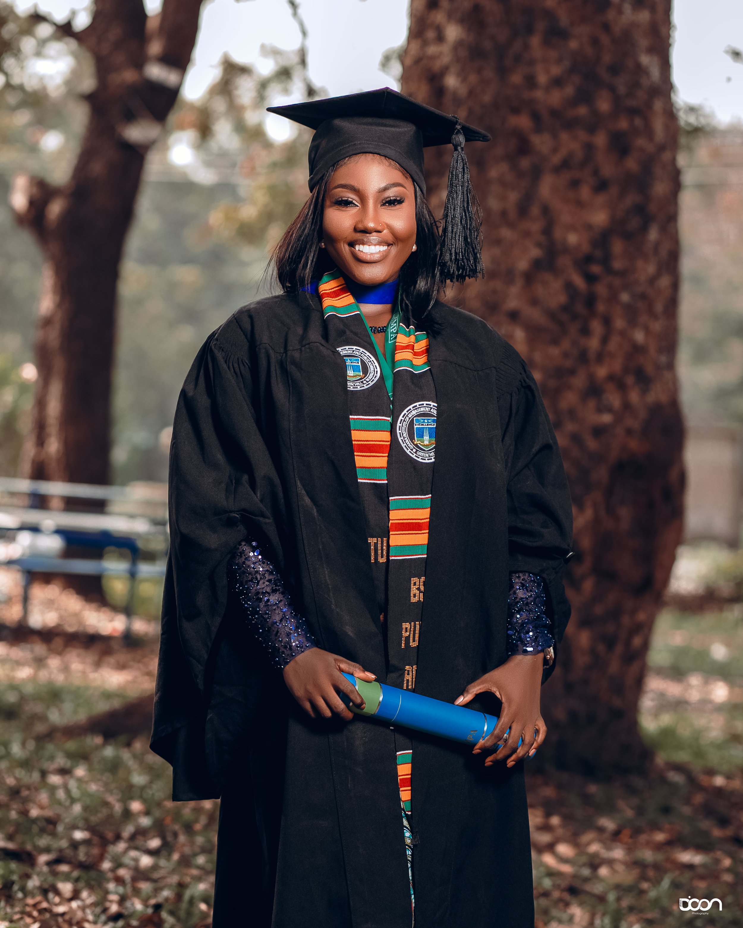 Young woman in graduation cap and gown holding diploma, outdoors with trees in the background.