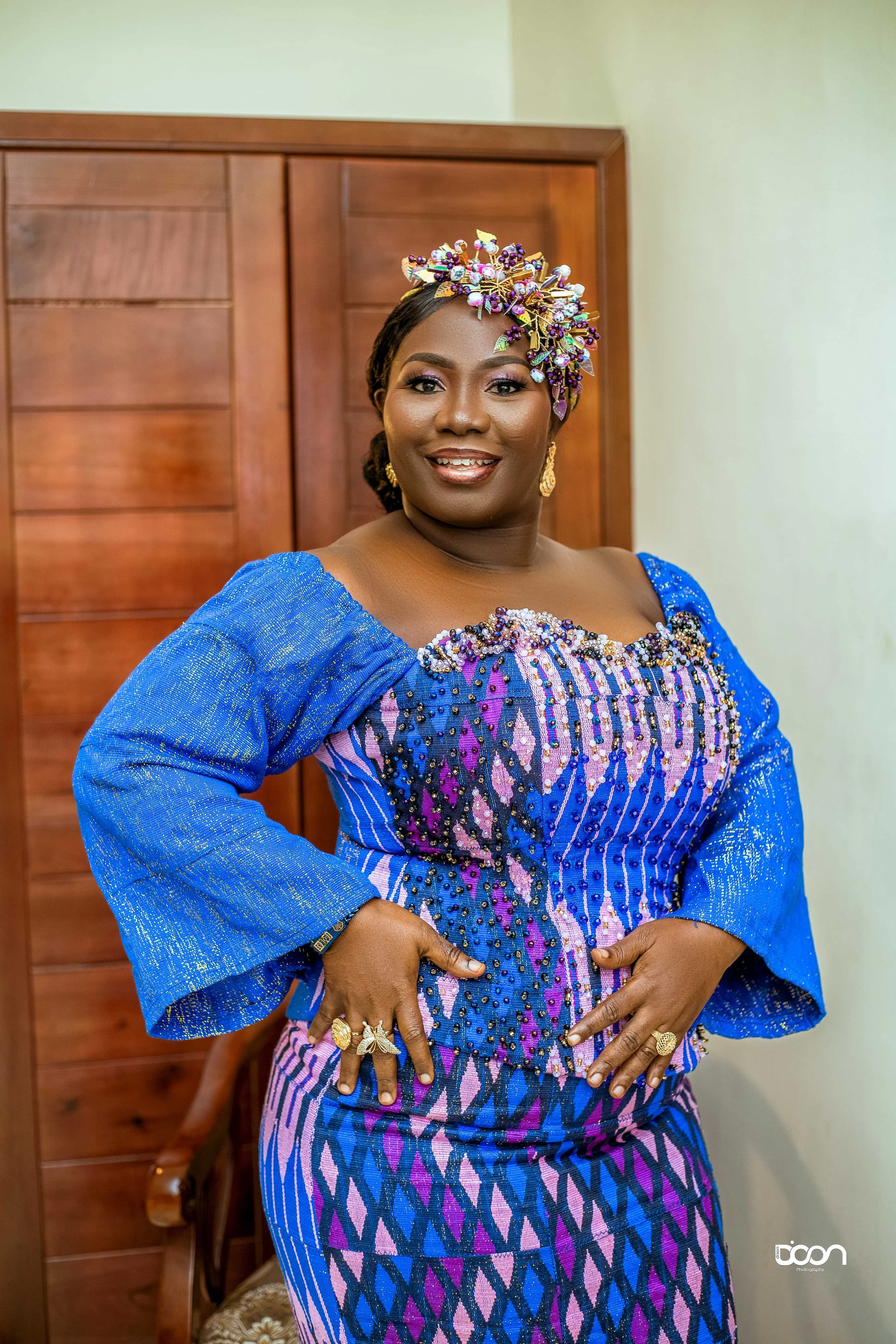 A woman dressed in a vibrant blue and purple traditional African attire, adorned with jewelry and an elaborate headpiece, smiling while standing indoors.