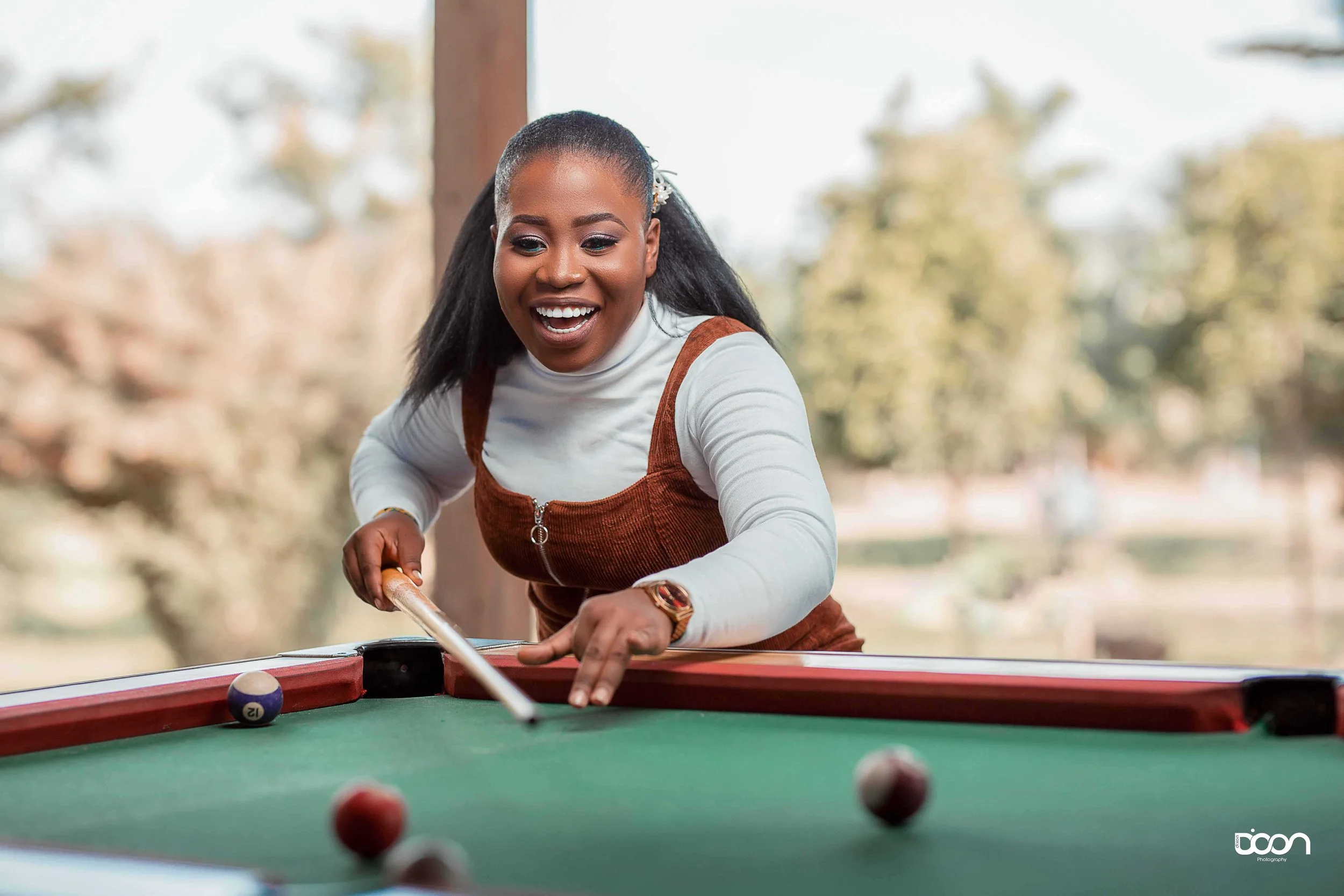 A woman playing pool, smiling, near a window with trees outside.