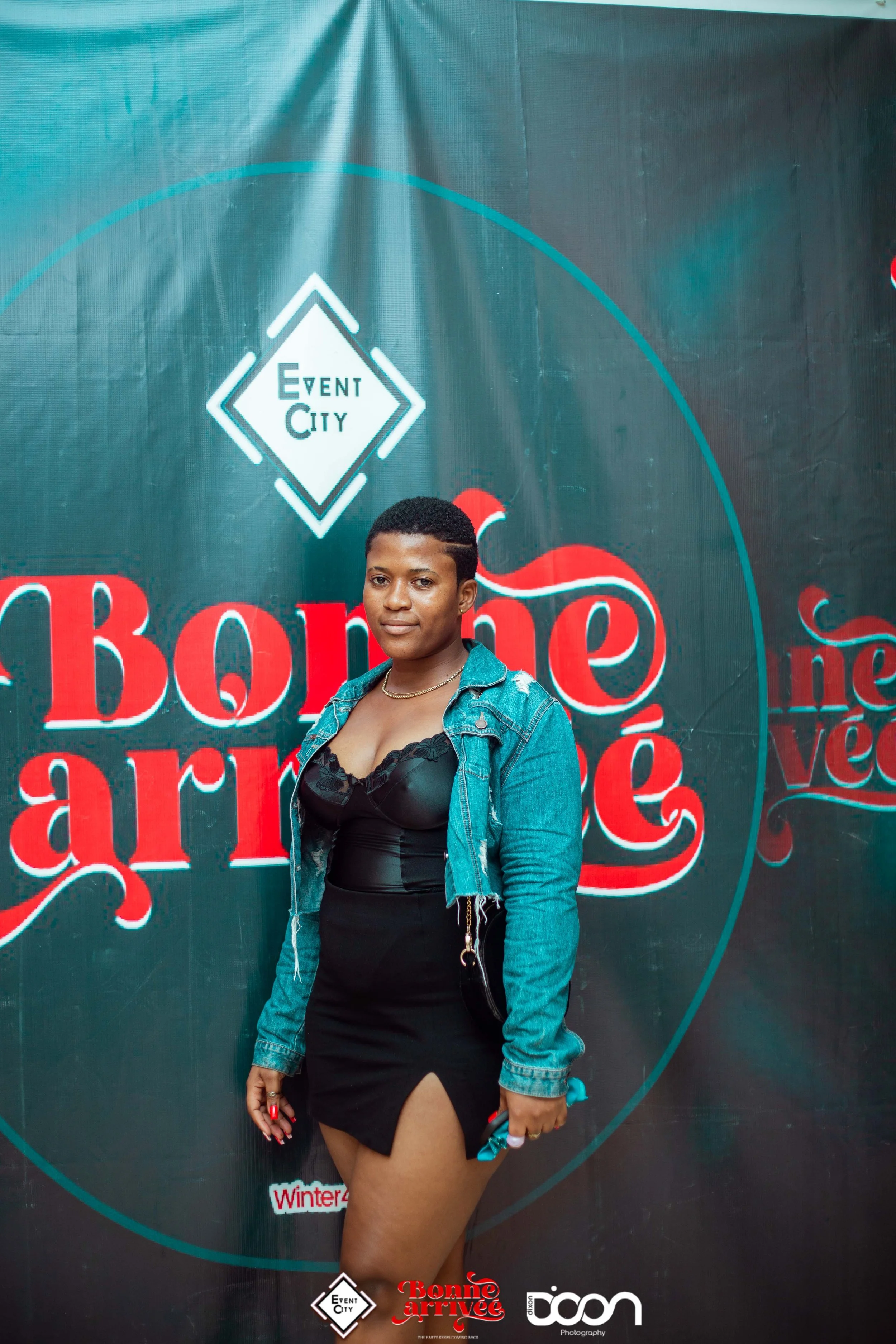 A woman standing in front of a dark background with red and white text, wearing a black dress with a slit and a denim jacket.