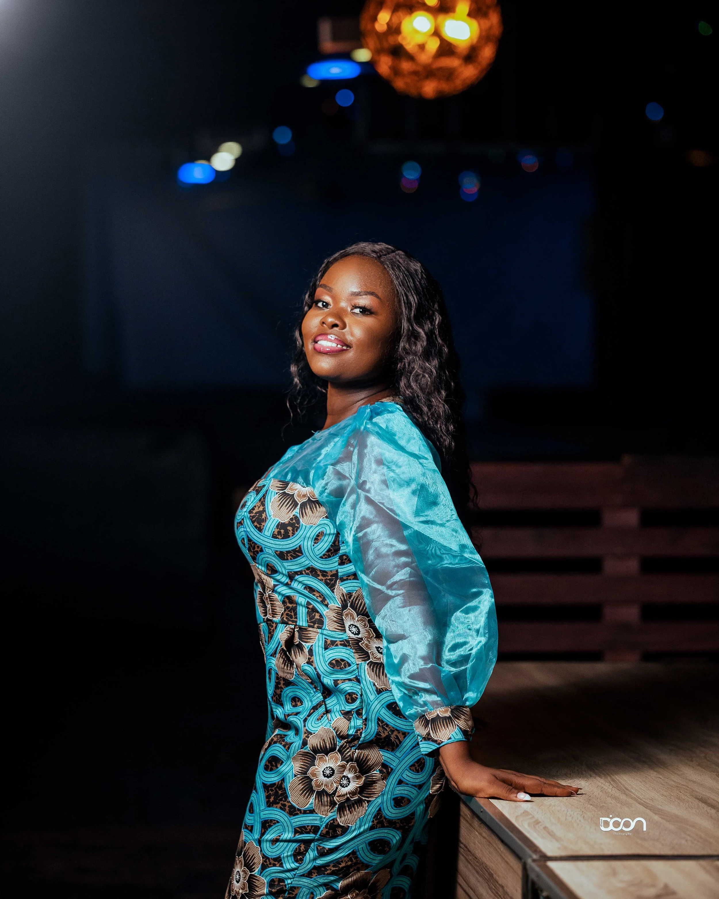 African American woman in colorful dress standing beside a wooden table in a dimly lit room with a decorative orange light fixture overhead.