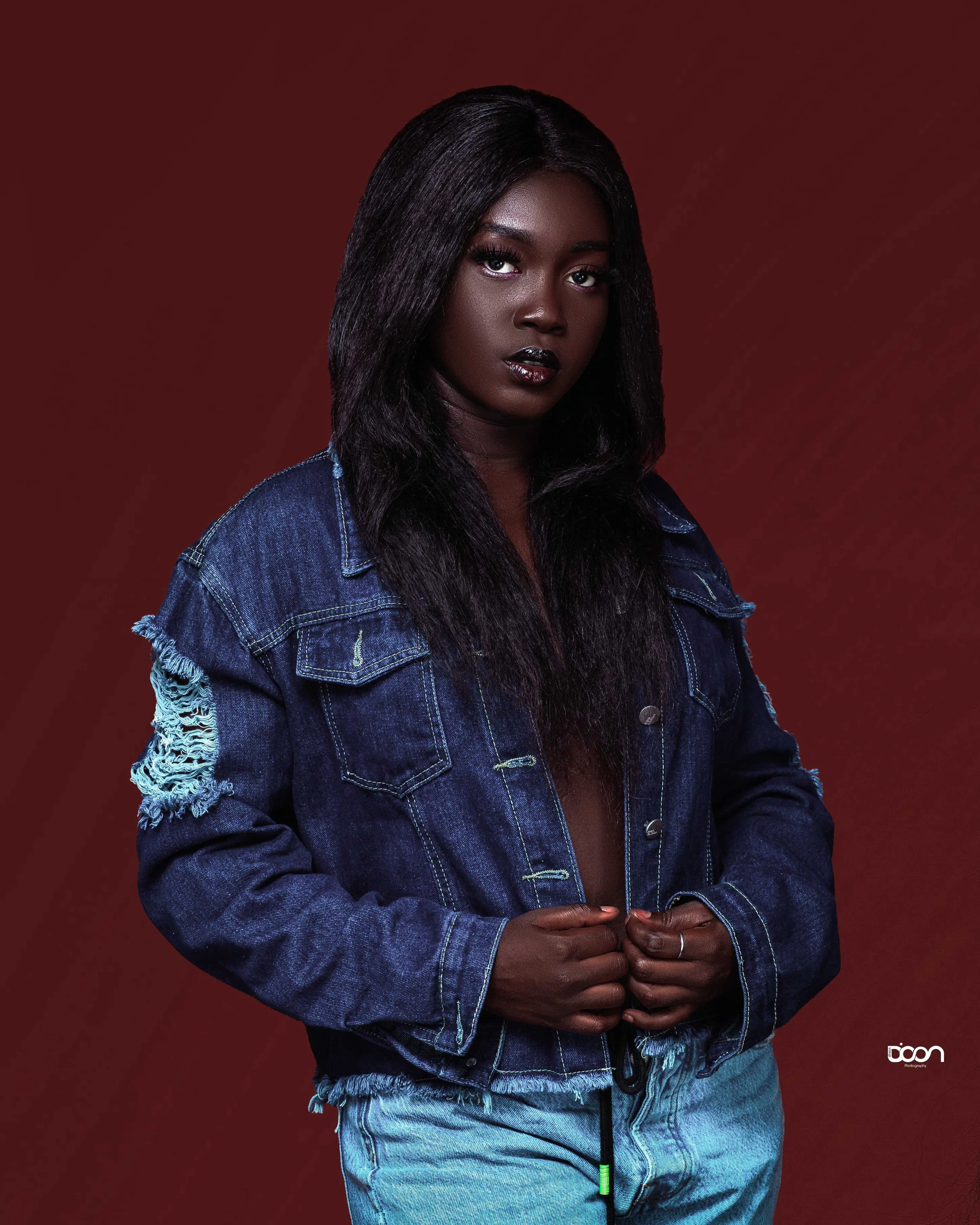 A young Black woman with long dark hair, wearing a distressed blue denim jacket and light blue jeans, standing against a dark red background. She has dark lipstick and a serious expression.