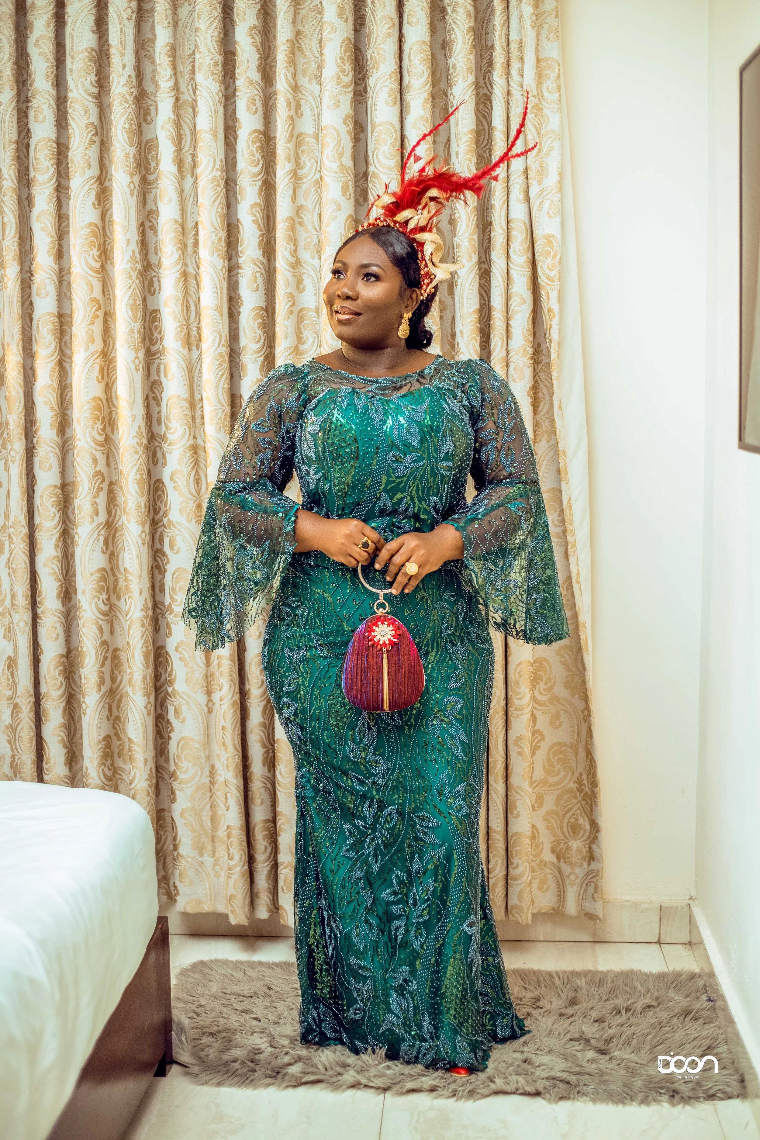 Woman wearing a green lace dress with sequins, holding a red beaded clutch, standing on a furry rug in front of beige patterned curtains, with a red feathered headpiece and gold jewelry, smiling.