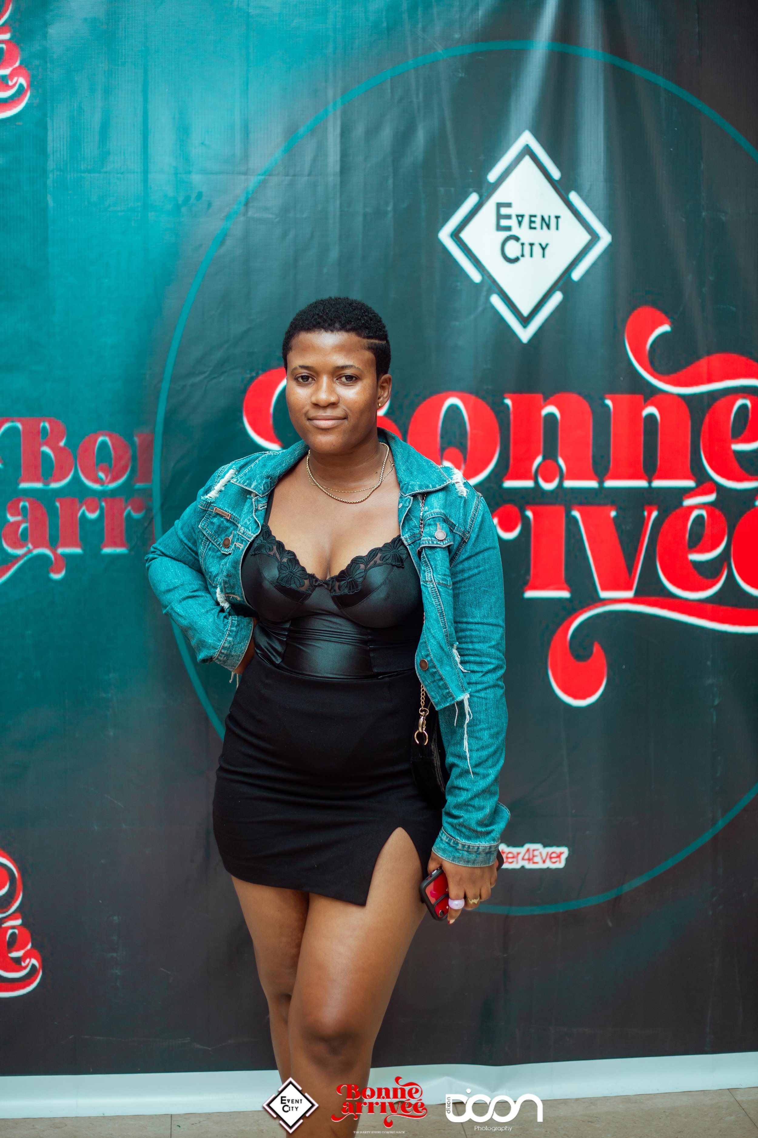 A woman standing in front of a backdrop with event logos, wearing a black dress with a thigh-high slit and a denim jacket, holding a phone in her right hand.
