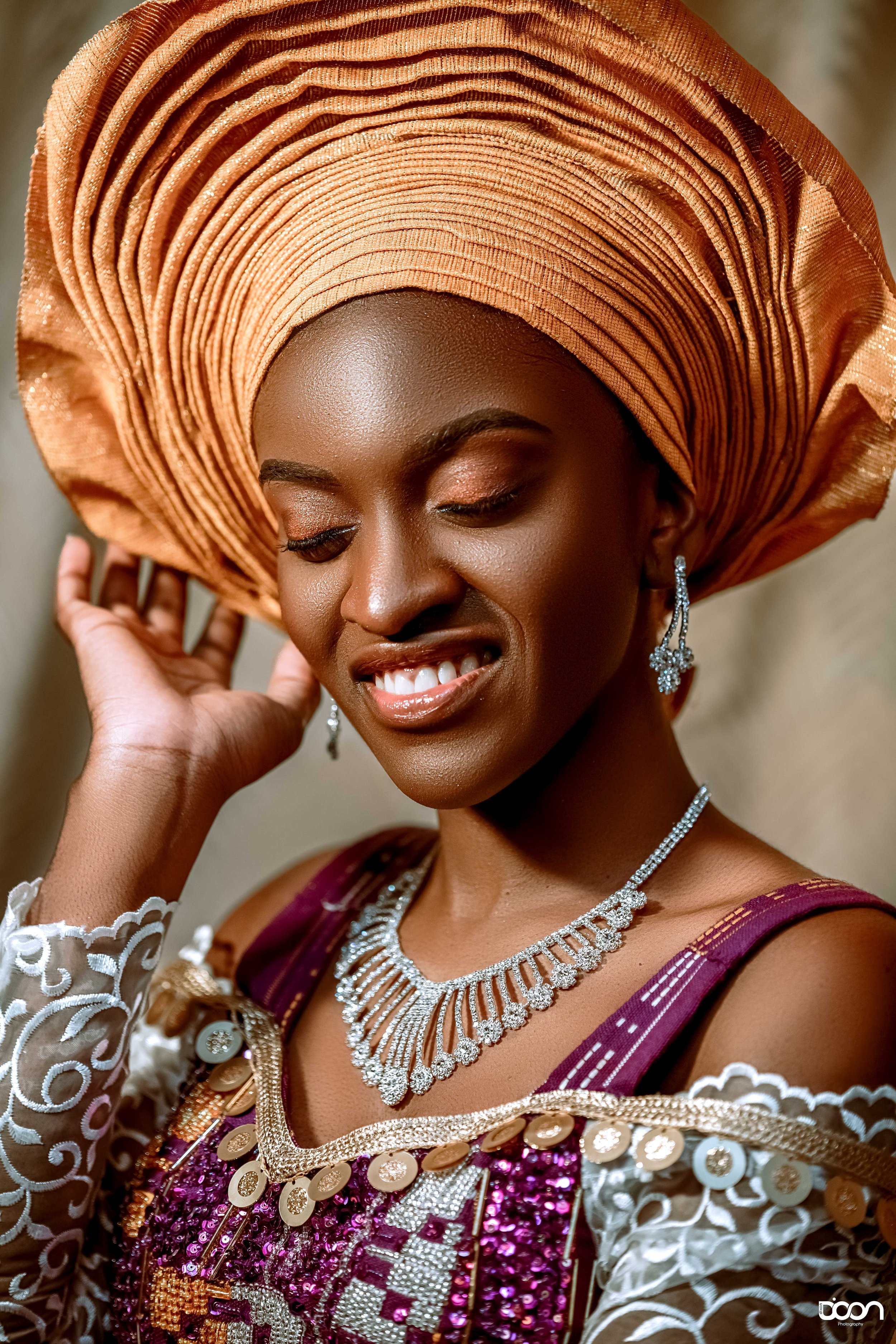 A woman dressed in traditional Nigerian attire, wearing an elaborate orange gele headwrap and a richly decorated burgundy and gold outfit, smiling with eyes closed and earrings, necklace, and lace sleeves visible.