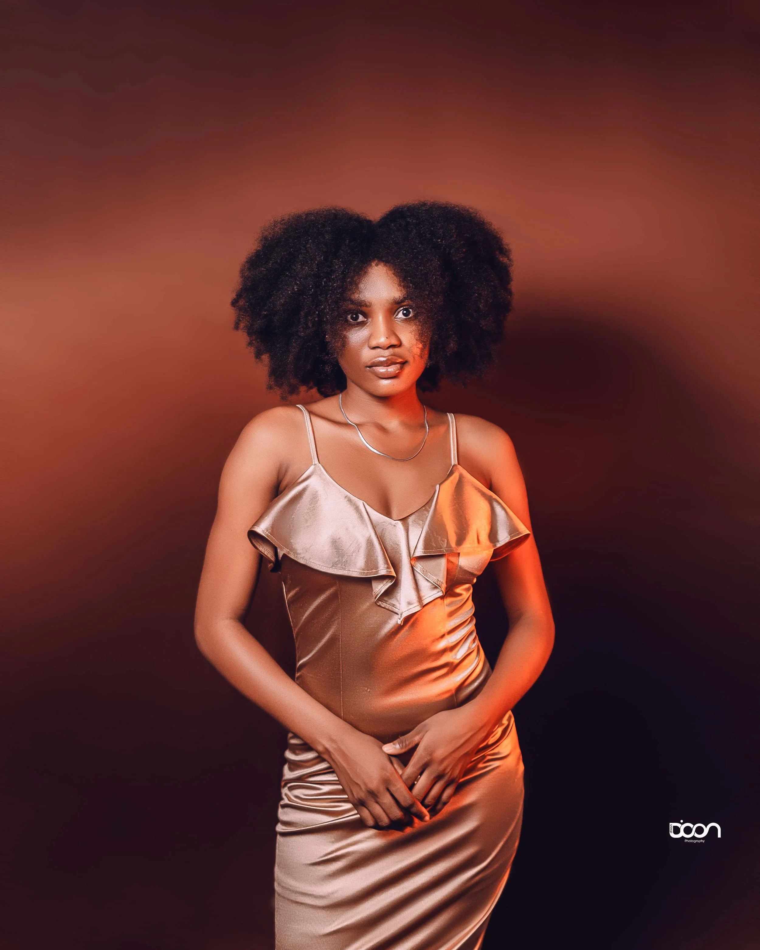 A woman wearing a shiny, satin gold dress with ruffles on the front, standing against a dark and warm-toned background with gradient shades of brown and orange. She has voluminous curly hair and is looking at the camera with a neutral expression.