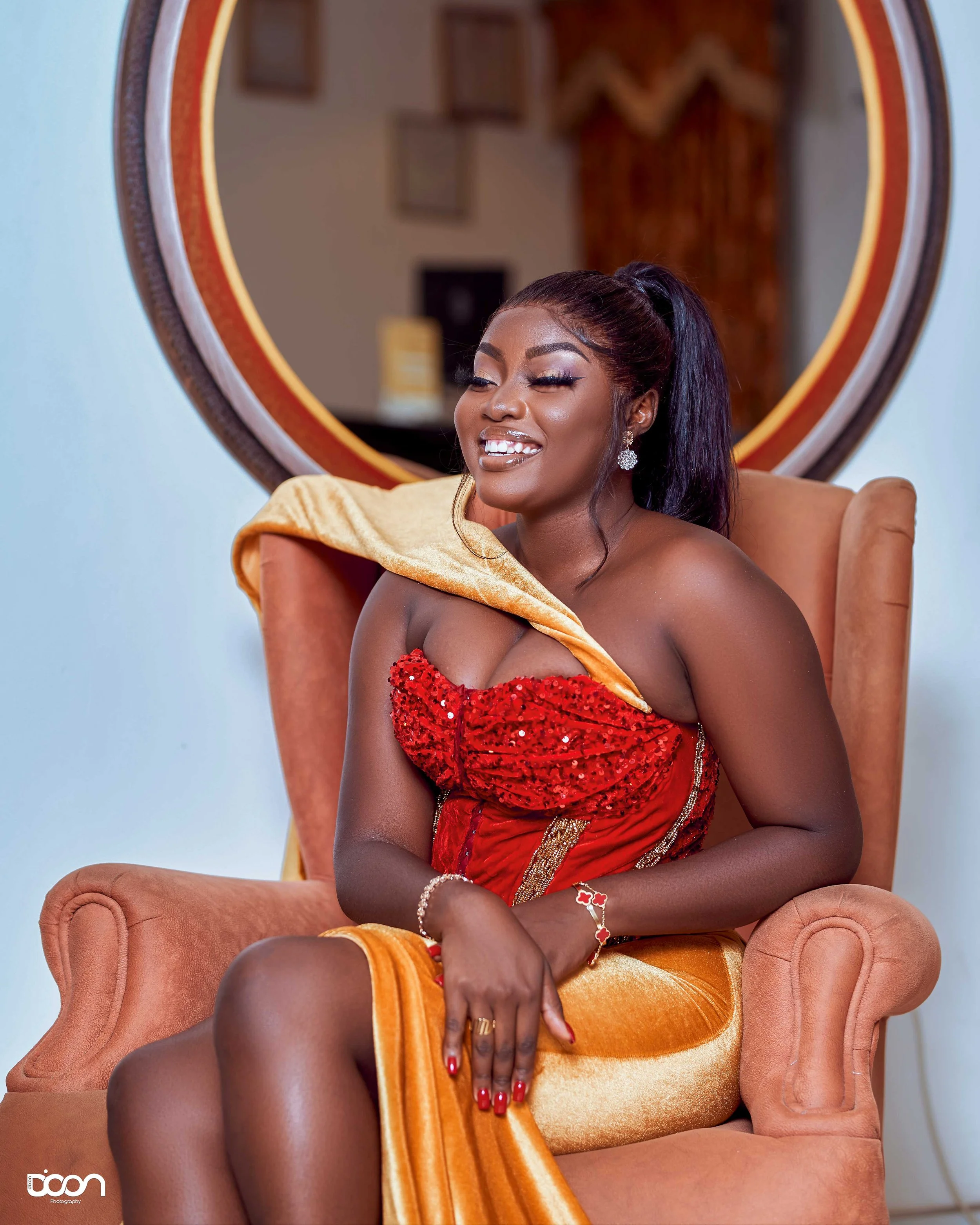 A woman sitting on an orange armchair, wearing a red sequin dress with a yellow shawl draped over her shoulder, smiling and posing for the camera with a mirror and framed pictures in the background.