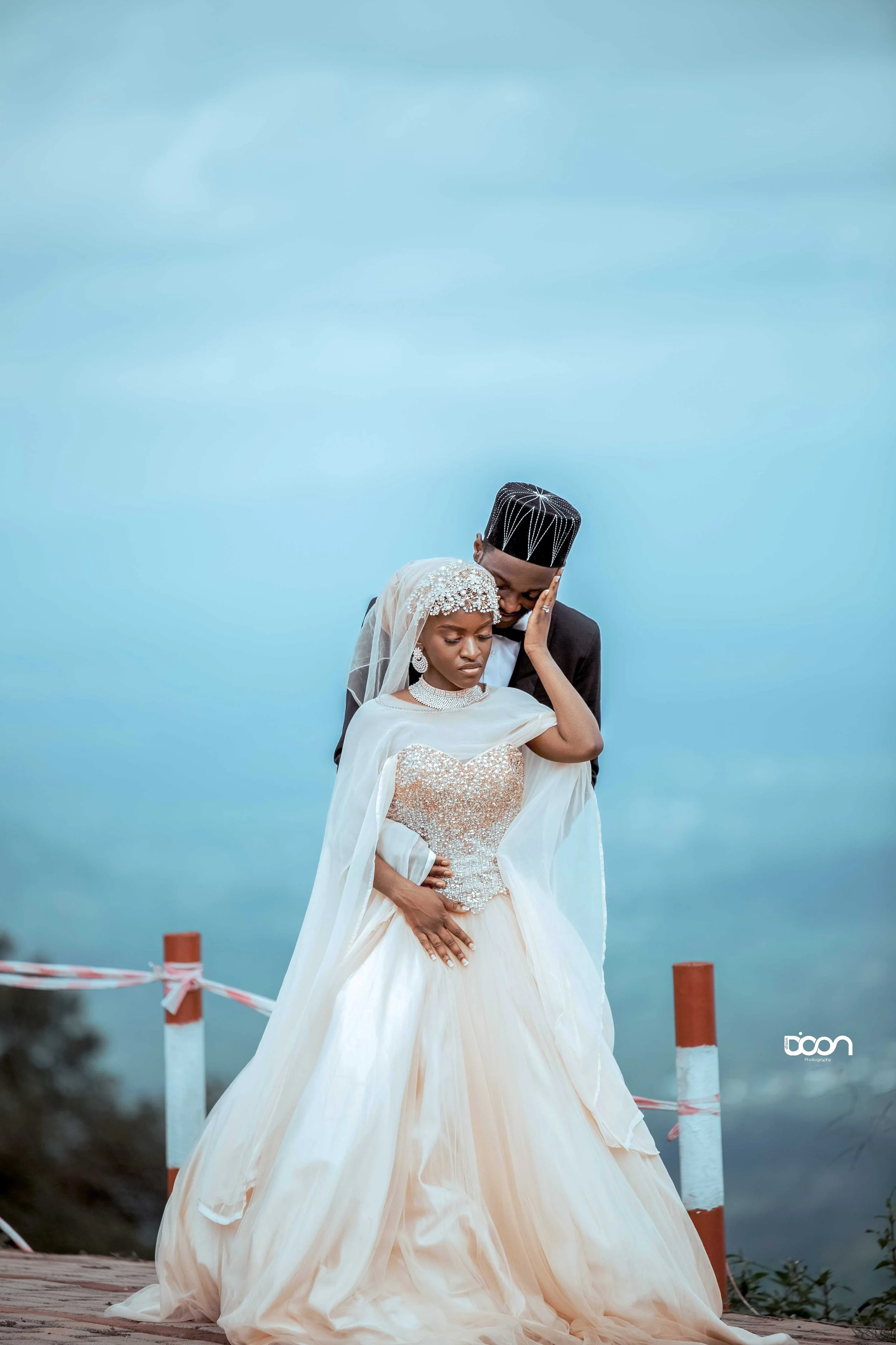 A bride and groom, dressed in traditional wedding attire, standing closely together outdoors with a blue sky and body of water in the background.