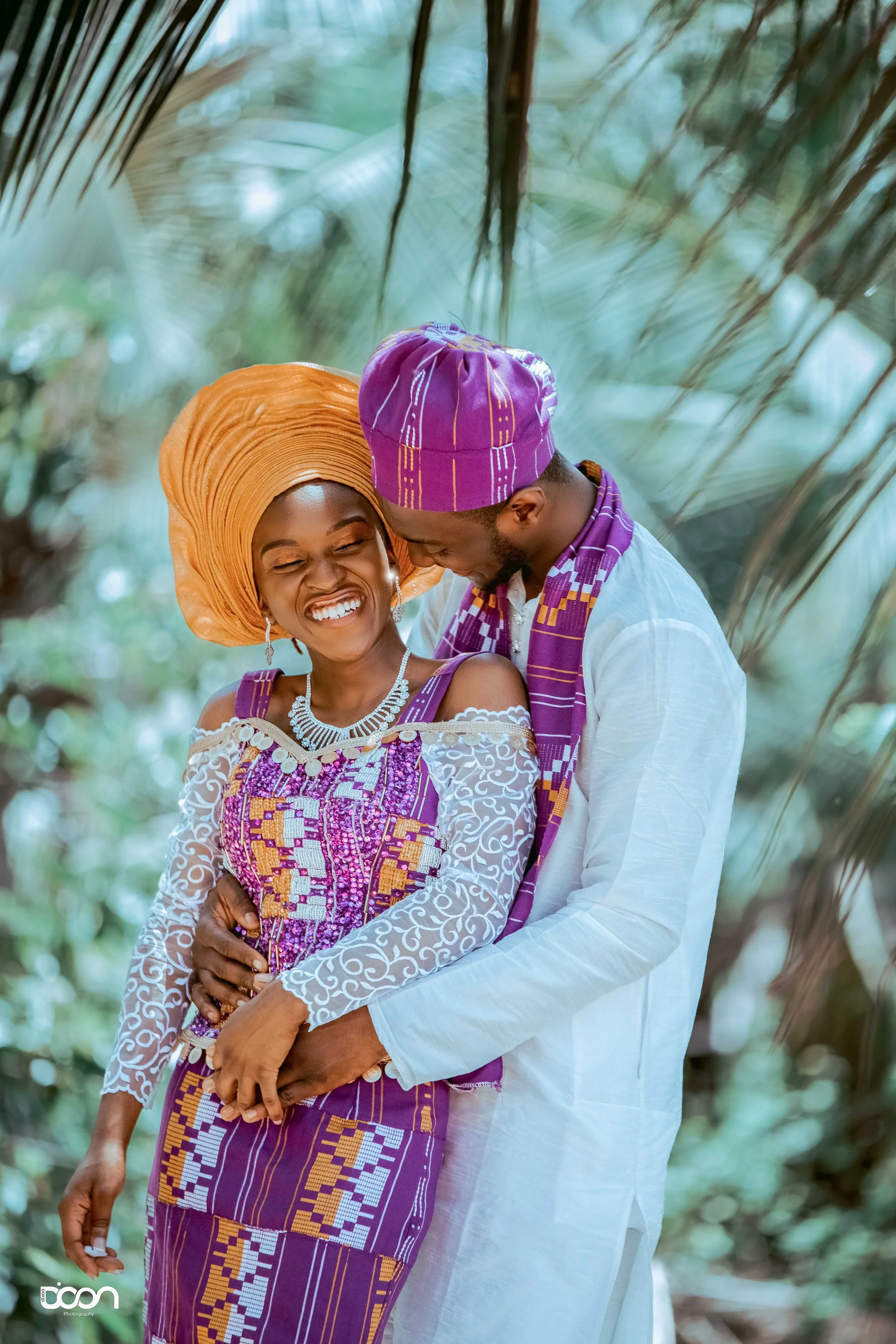 A joyful couple dressed in traditional African attire, smiling and embracing outdoors among lush greenery.